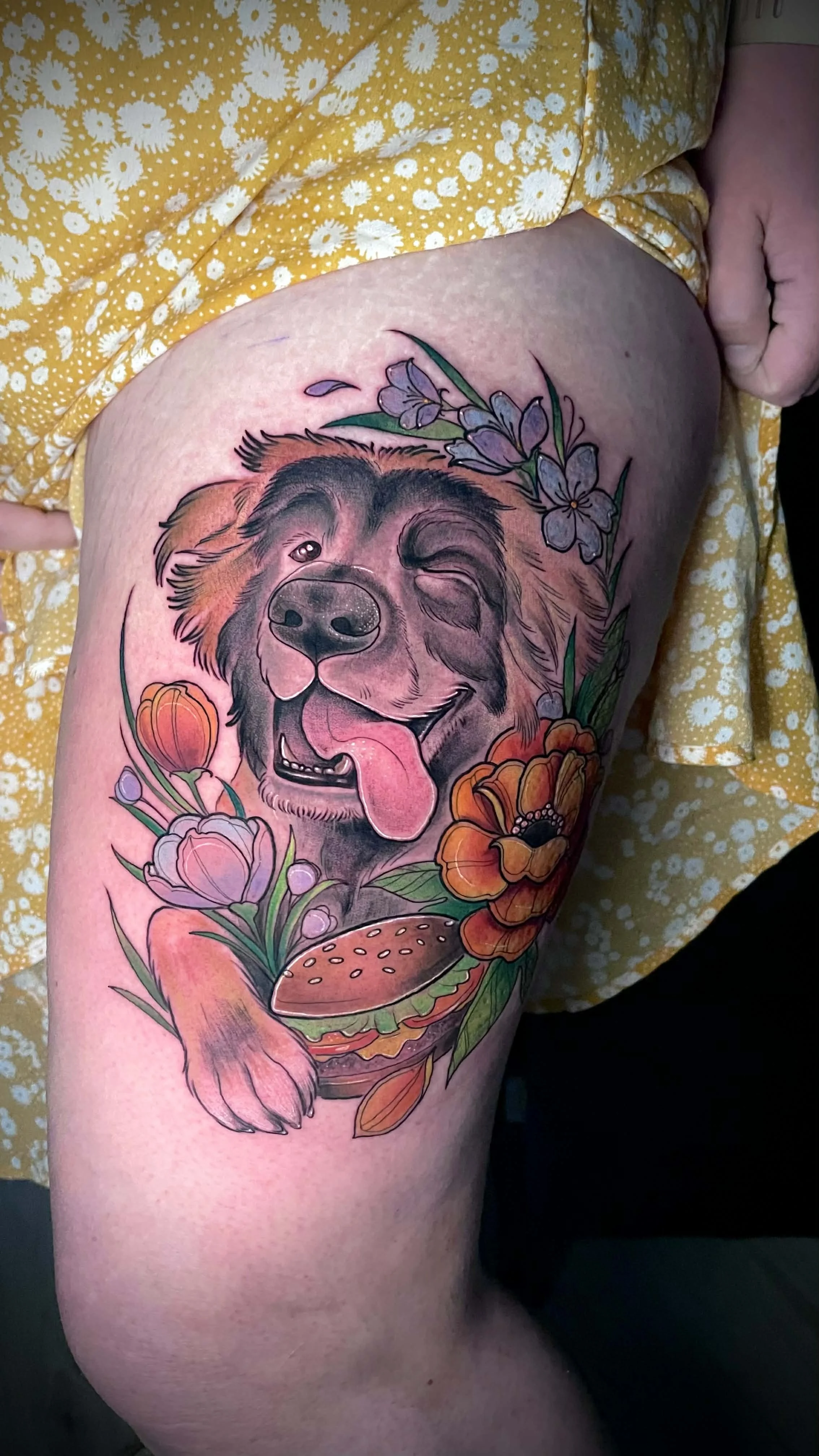 Tattoo of a happy, winking dog with its tongue out, surrounded by colorful flowers and a burger on the thigh.