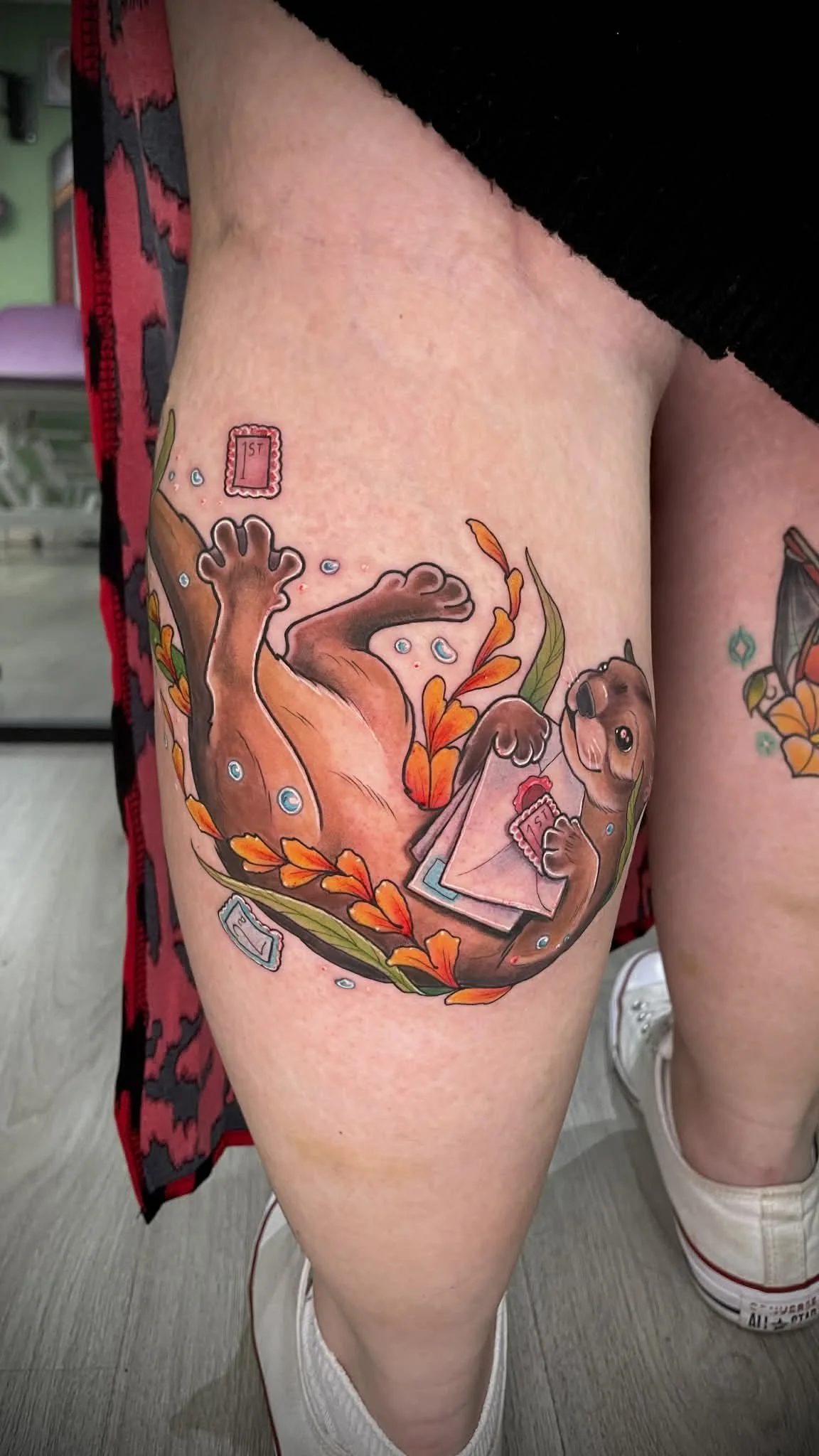 Tattoo of an otter holding an envelope with a pink stamp, surrounded by orange and red leaves on a person's calf.