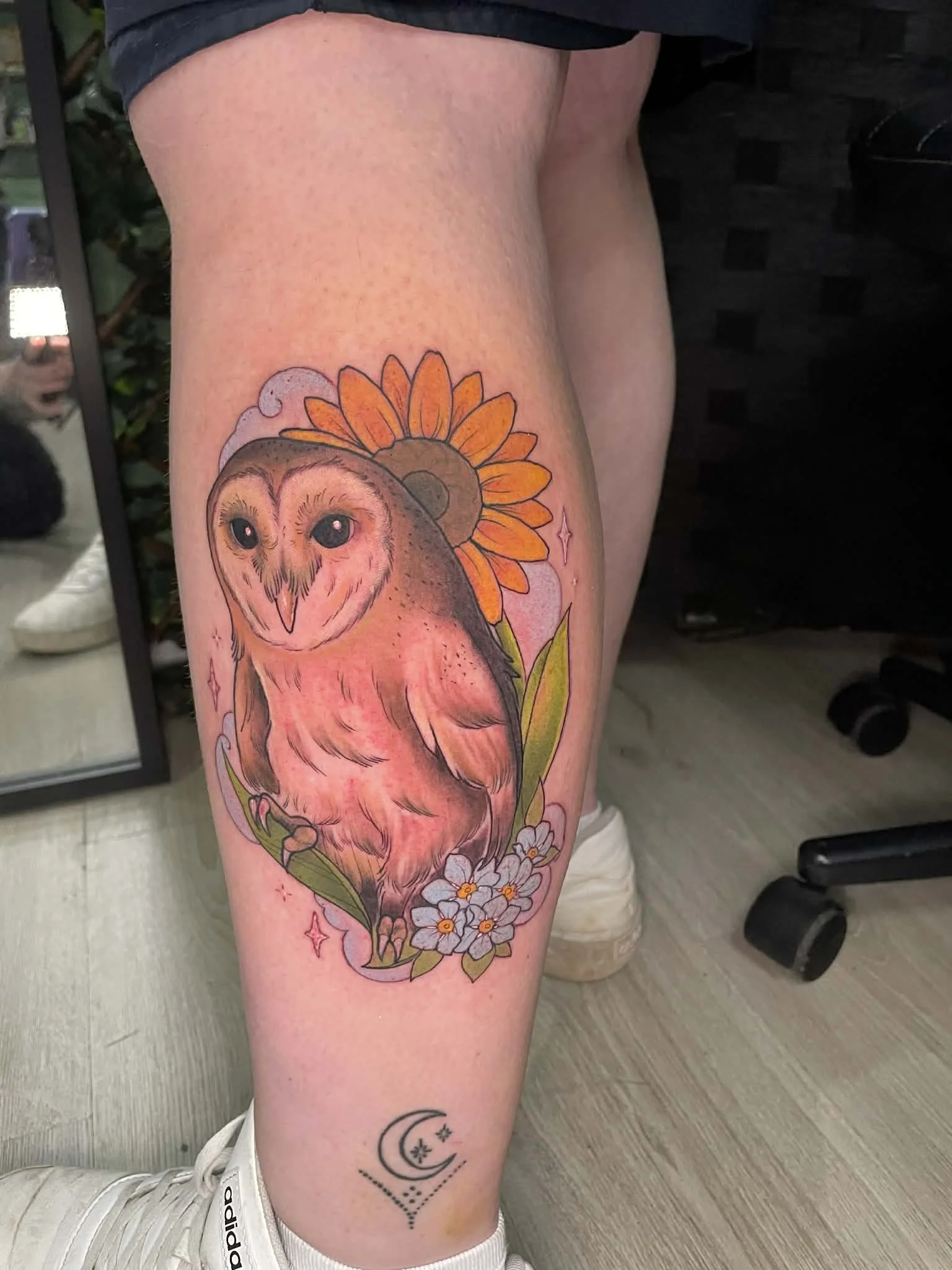 Tattoo of an owl with a sunflower behind it, surrounded by smaller flowers and sparkles, on a person's thigh.