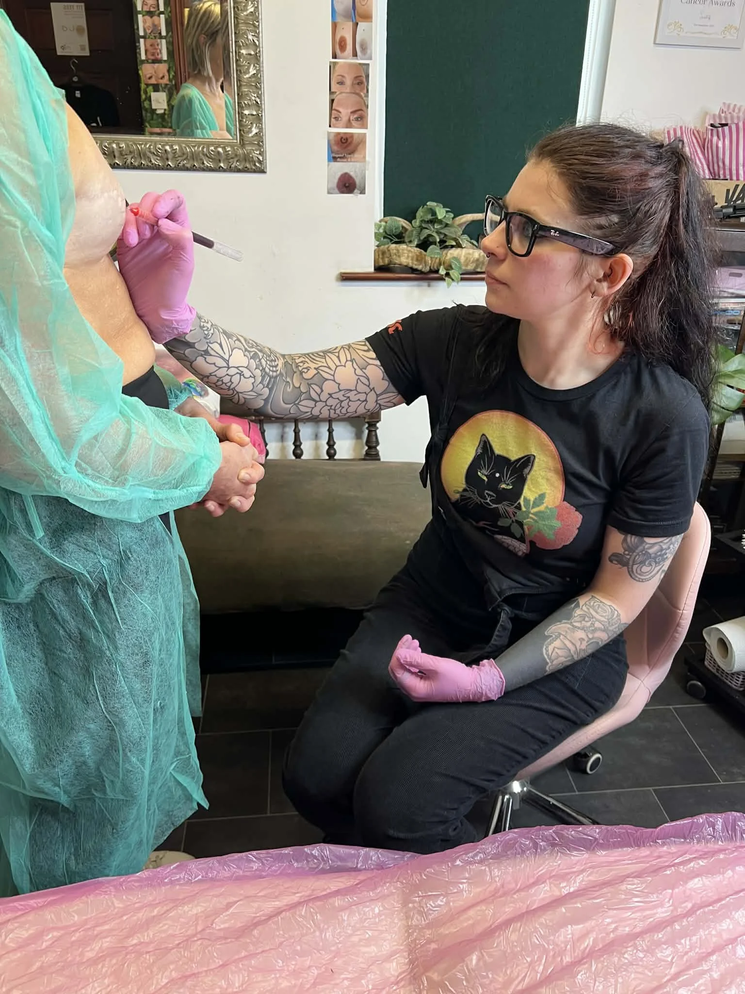 Tattoo artist with glasses and black T-shirt sitting on a pink chair, getting ready to draw or tattoo on a person's chest; tattoo artist has floral tattoos on her arms and wears pink gloves, and the person receiving the tattoo is covered with a green disposable gown. There are photos on the wall, a mirror, and plants in the background.