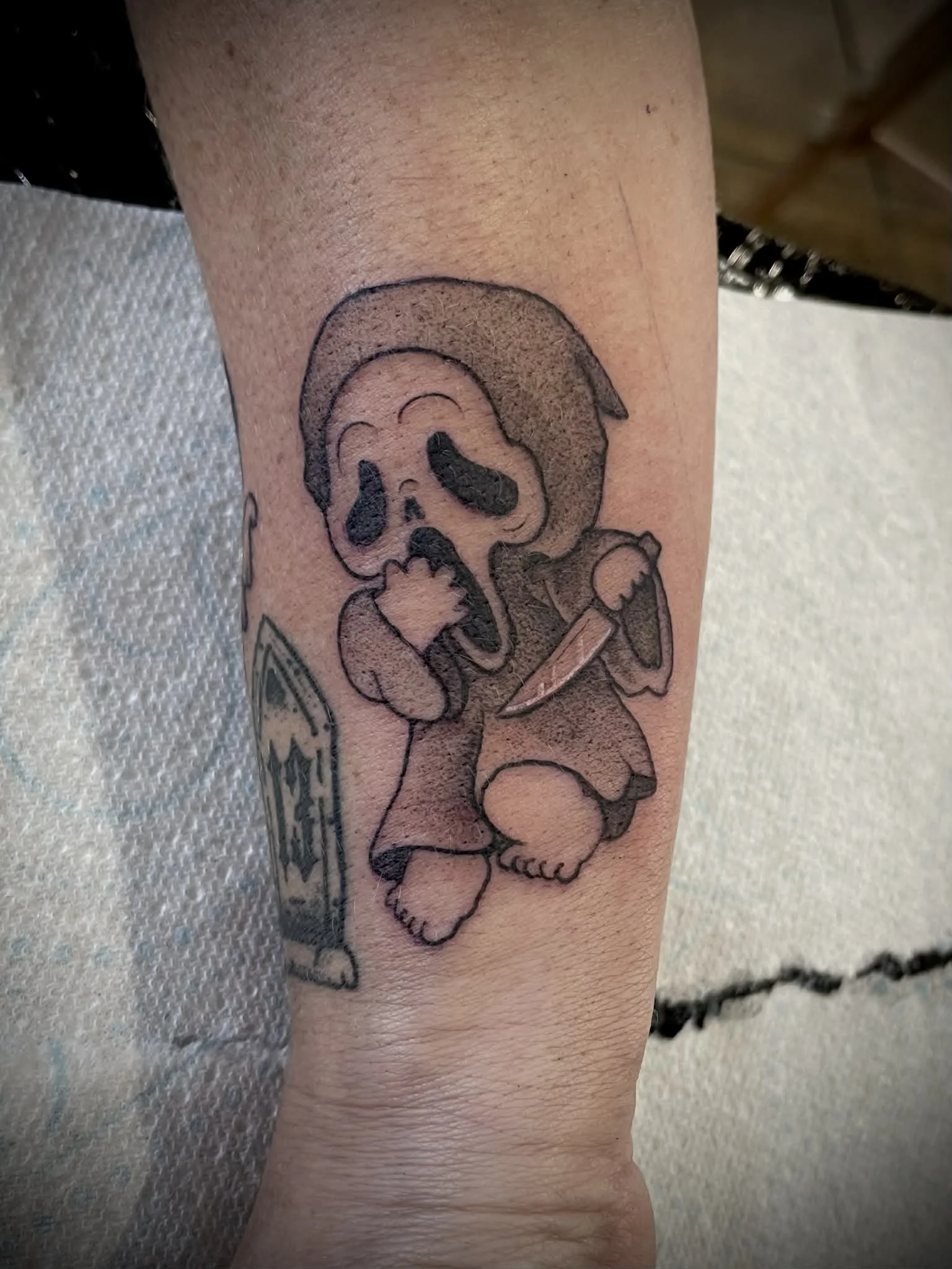 Tattoo of a cartoon skull character with a large head, dark eye sockets, and a tongue sticking out, holding a knife, with a skateboard nearby.