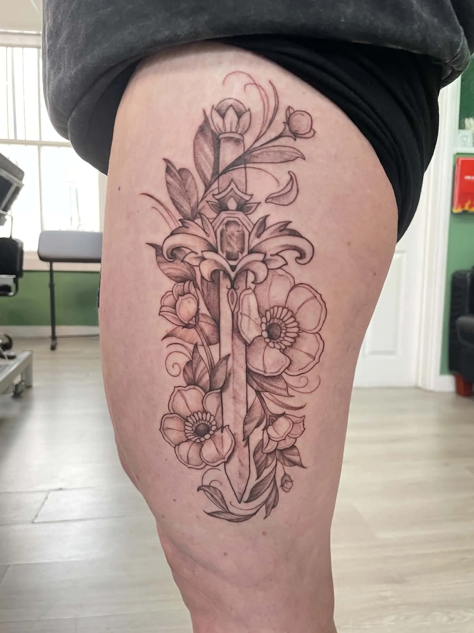 A detailed black and gray floral tattoo on a person's upper thigh, featuring various flowers, leaves, and intricate line work.