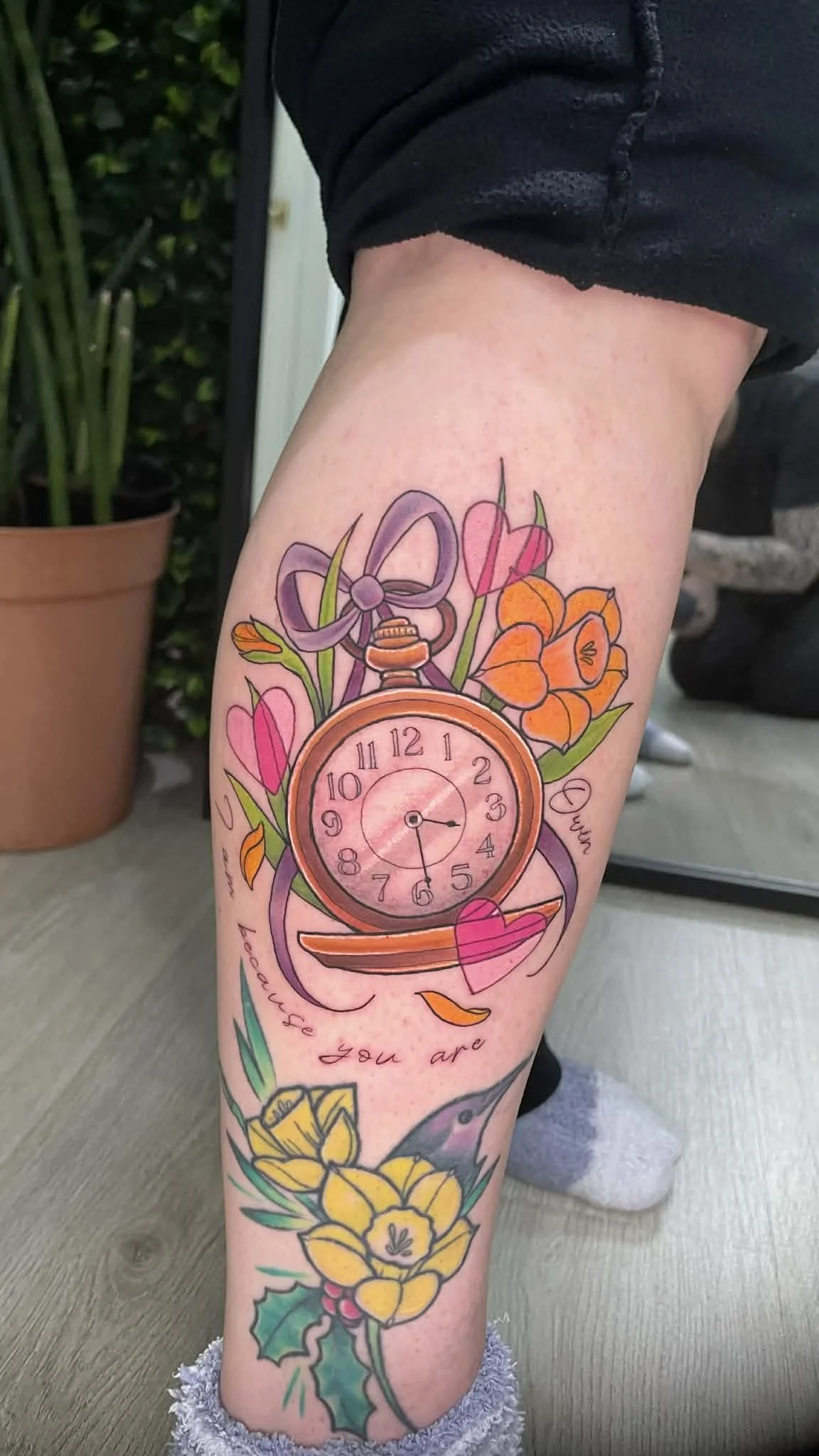 Colorful tattoo on a person's leg depicting a vintage alarm clock surrounded by flowers, a bow, a heart, and a hummingbird with cursive text.