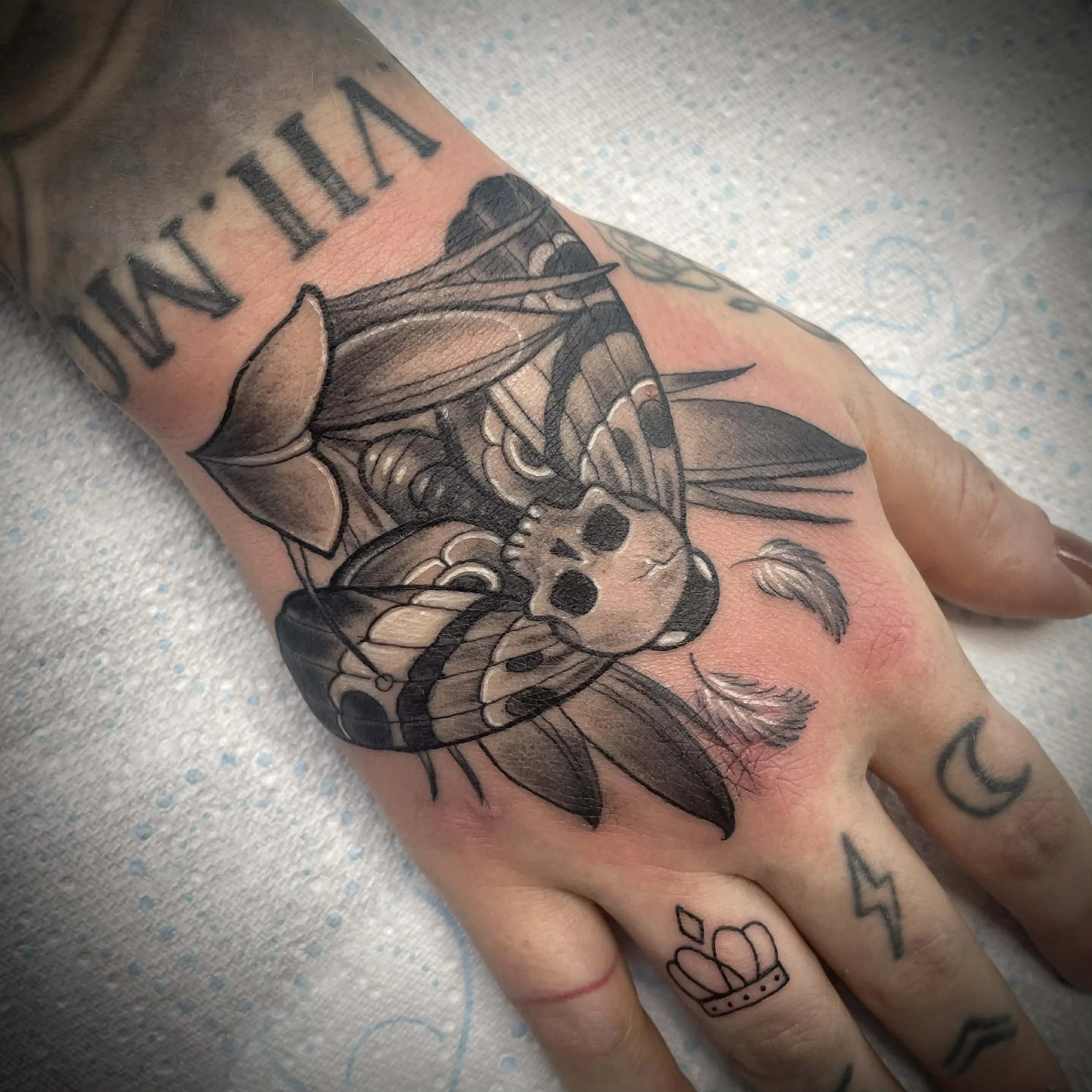 Black tattoo of a moth, skull, dandelion, and leaves on a hand with other smaller tattoos including a moon, lightning bolt, and crown.
