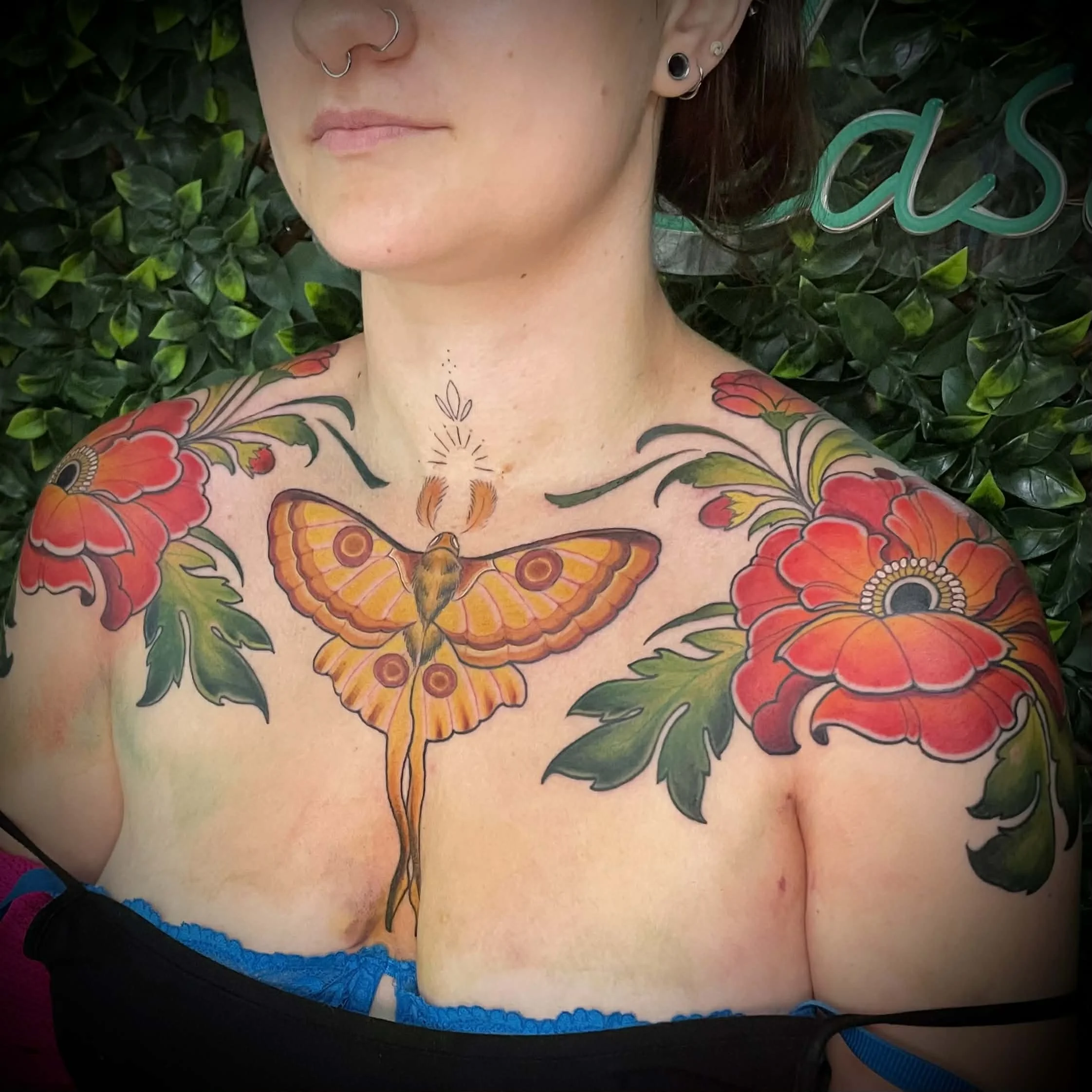 Colorful tattoo of a orange and yellow moth with intricate wing patterns, floral designs in red and green, on a person's chest and shoulders, with greenery in the background.