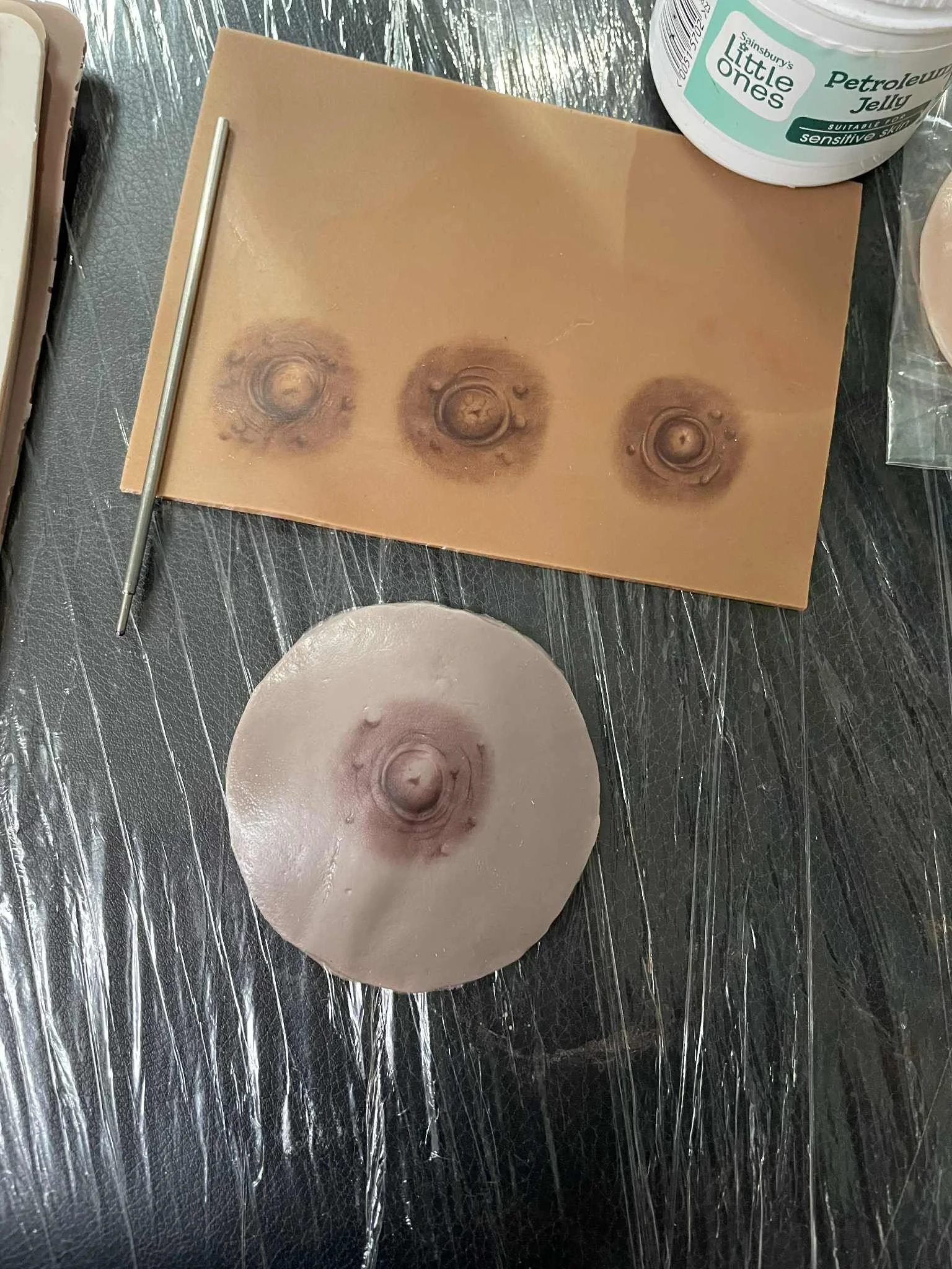 A silicone mold with three circular indentations, and a circular silicone piece with a similar indentation, on a black surface. A metal tool is placed beside the mold, and a container of Sainsbury's Little Ones Petrelow Jelly is partially visible in the upper right corner.