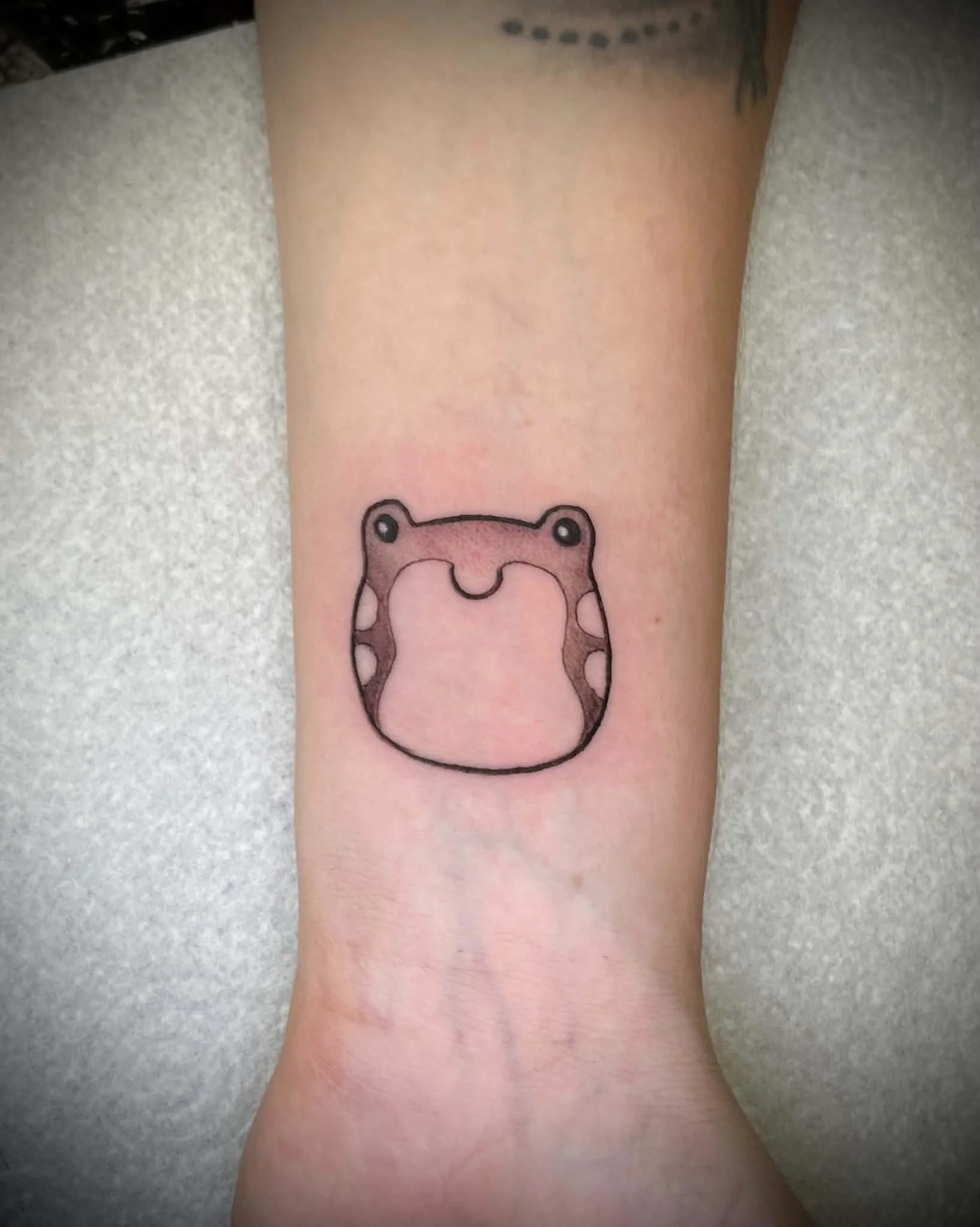 A person's arm with a tattoo of a cute cartoon animal face, possibly a bear, featuring a pink nose, black eyes, and a smiling mouth, on the inner forearm.