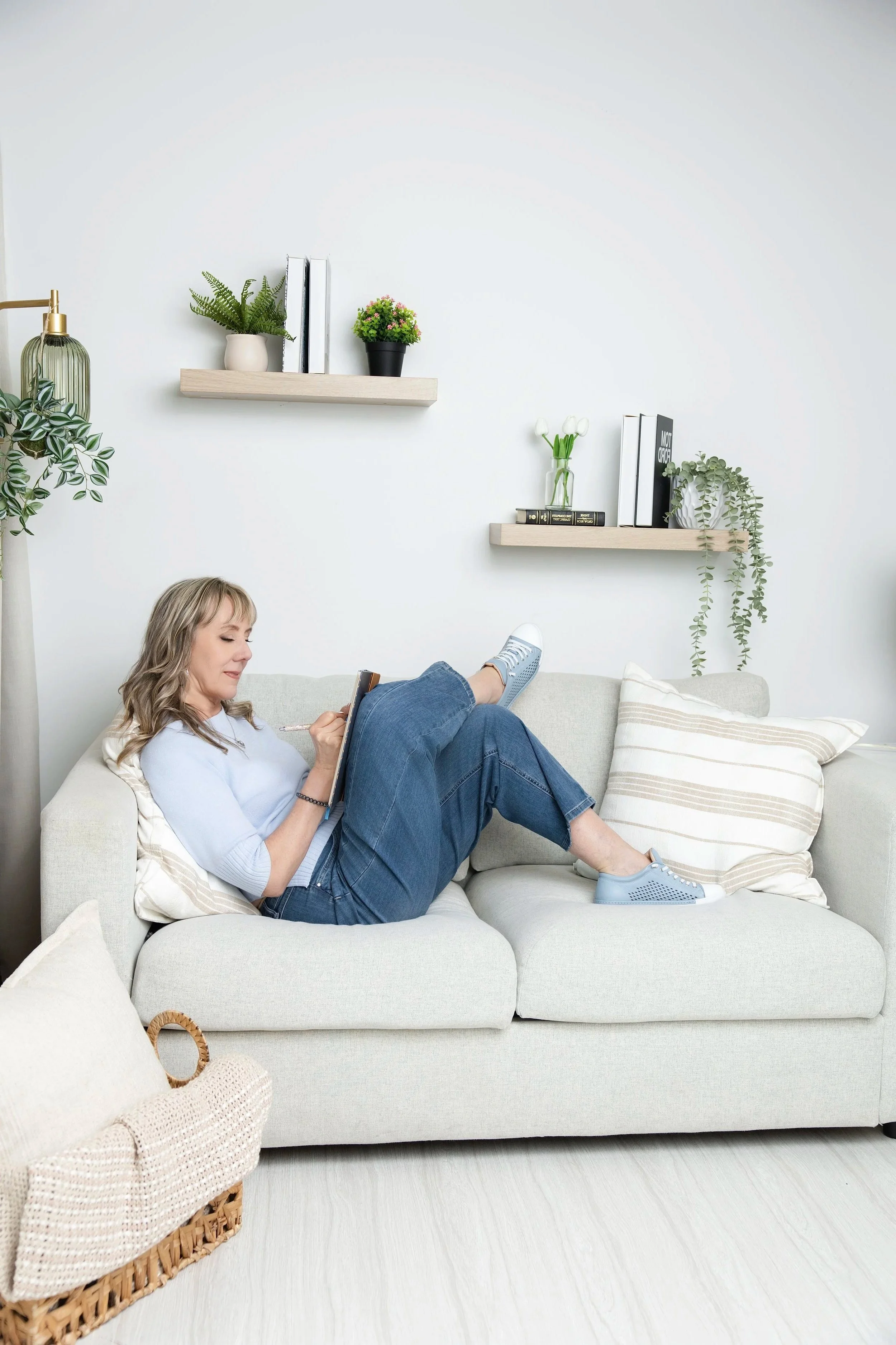A woman with blonde hair sitting on a light-colored sofa, relaxing with her legs up, wearing a light blue shirt, jeans, and sneakers, in a bright, minimally decorated living room with floating shelves, plants, and books.