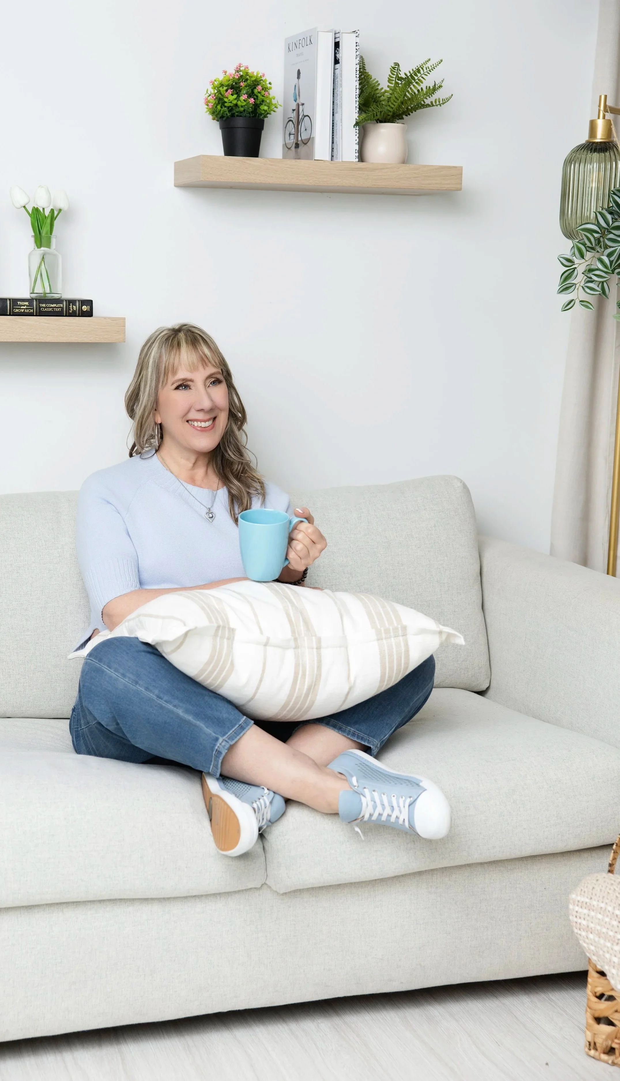 A woman sitting on a light-colored sofa, holding a blue mug, with a pillow on her lap, wearing a light blue shirt, jeans, and sneakers, in a cozy living room with shelves, books, and potted plants on the wall.