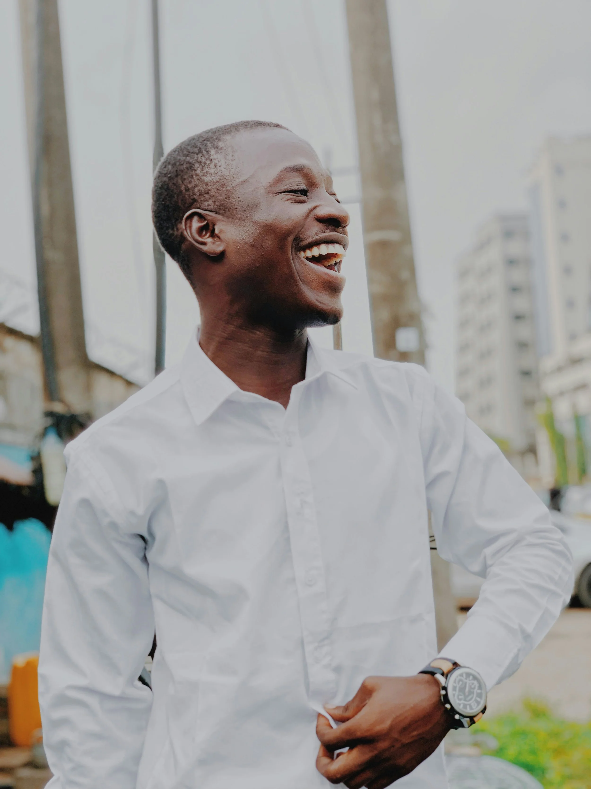 A young man with short hair wearing a white shirt and a watch on his wrist, smiling and looking to the left, standing outdoors with buildings and poles in the background.