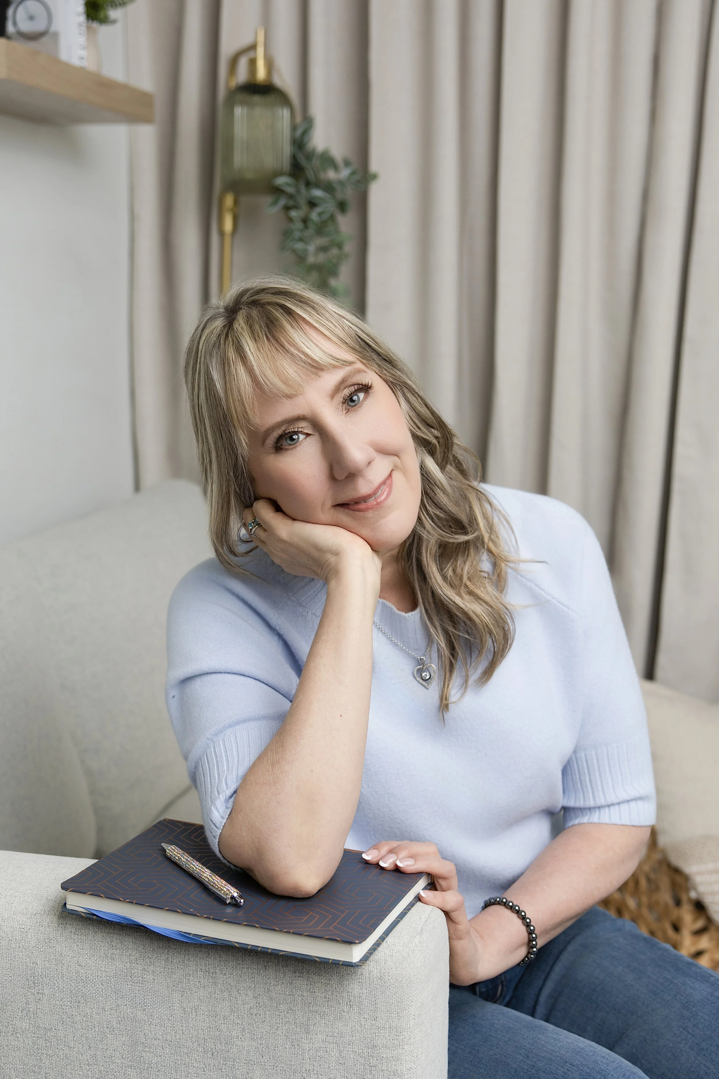A woman with blonde hair and blue eyes sitting at a table, resting her head on her hand, smiling slightly, with a notebook and pen on the table in front of her. She is wearing a light blue top and a heart-shaped necklace. The background features a beige curtain, a green plant, and a wall-mounted light fixture.