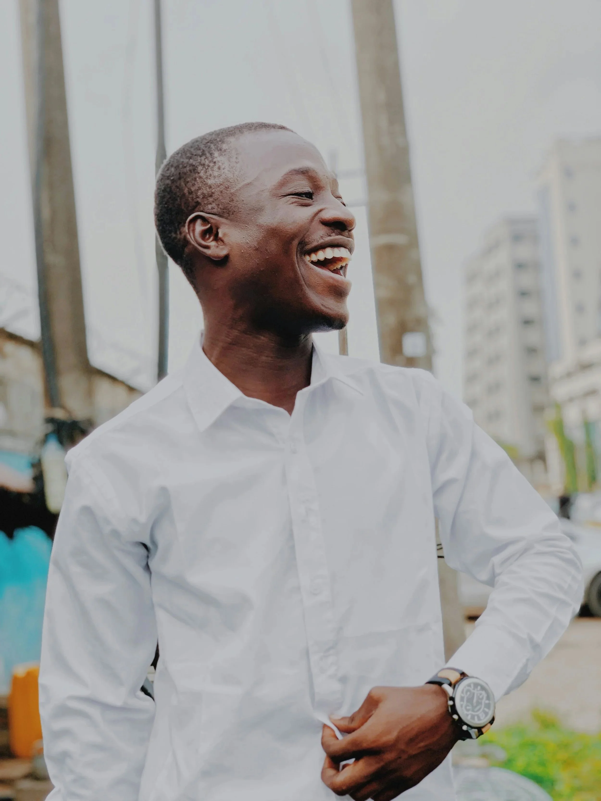 A man in a white button-up shirt smiling and laughing outdoors, with buildings and utility poles in the background.