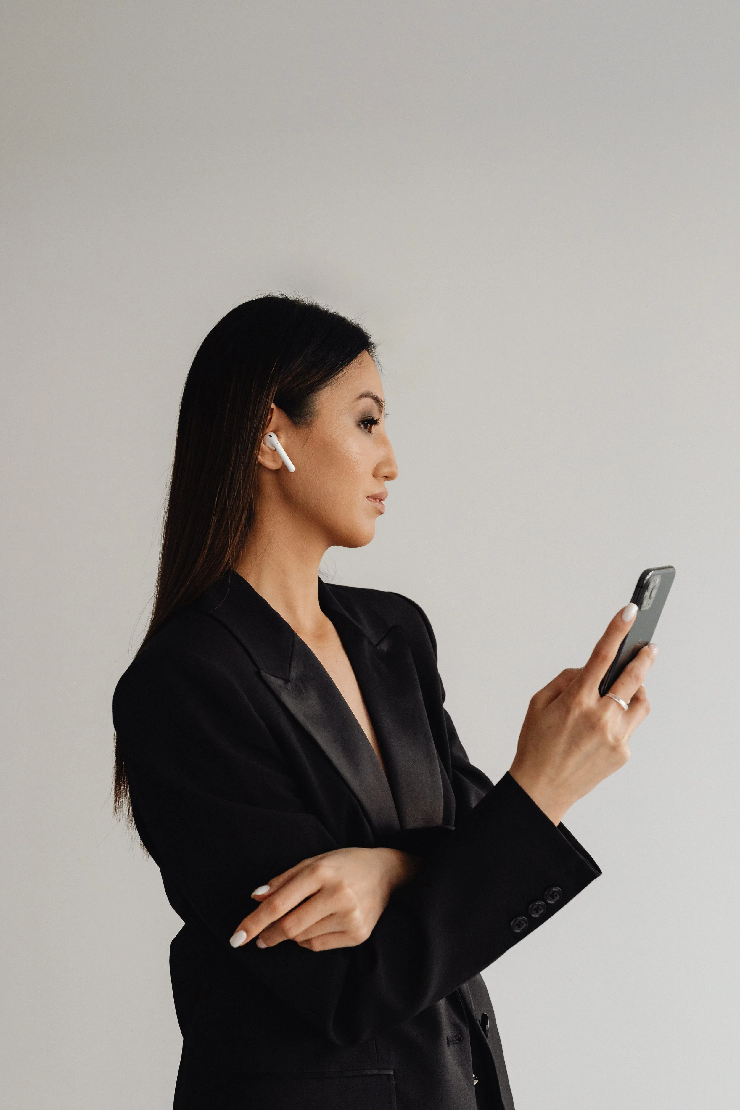 A professional woman with long dark hair is wearing a black blazer and white earbuds while looking at her phone. She has crossed arms and is standing against a plain light-colored wall.