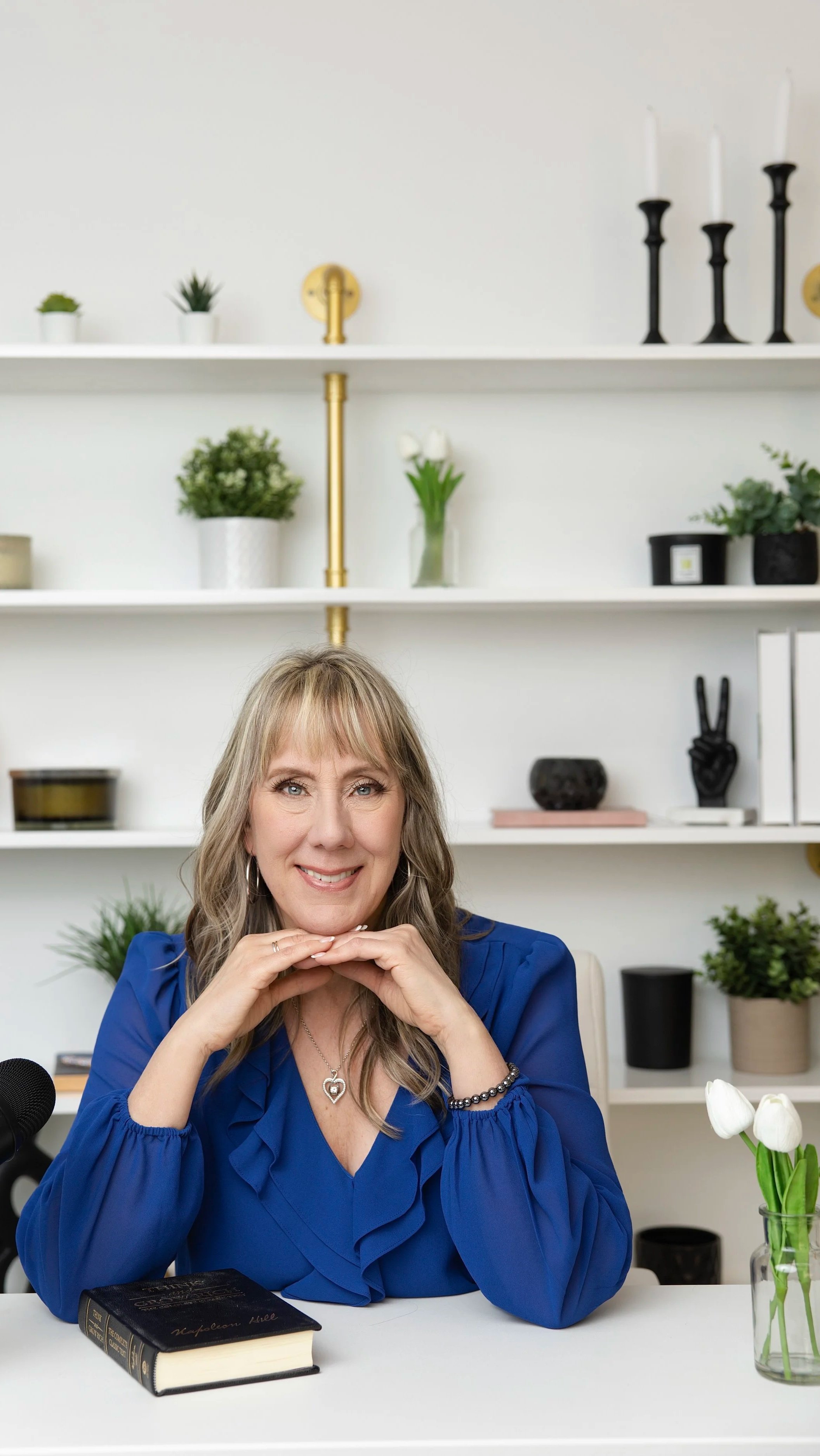A woman with long blonde hair in a blue blouse sitting at a white desk in a room with white shelves decorated with green plants, white tulips, black candlesticks, and books.