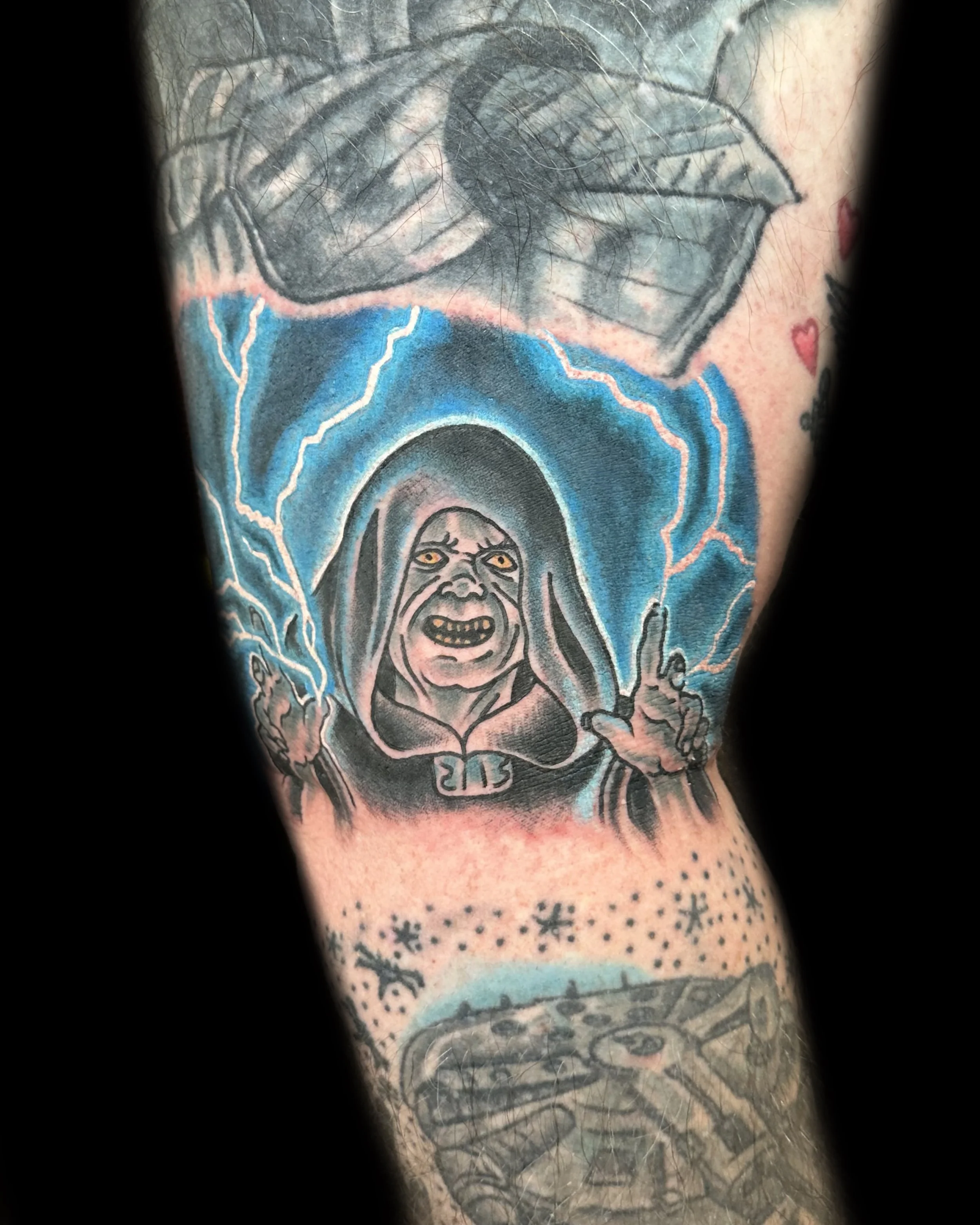 Tattoo of a woman with a hood and yellow eyes, surrounded by blue lightning, pointing with both hands.