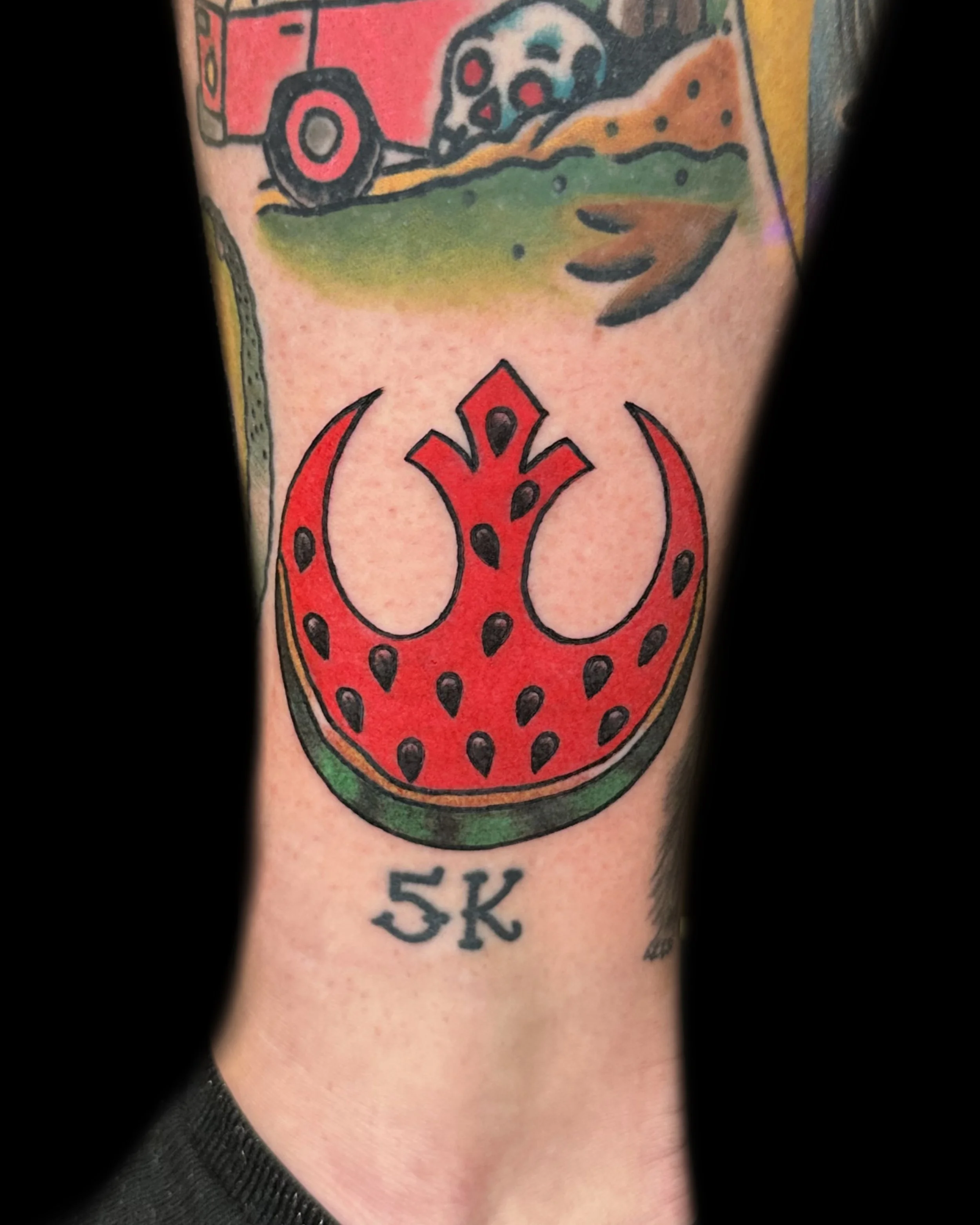 A colorful tattoo of a stylized red and black watermelon slice with a green rind, with the text "5K" below it.