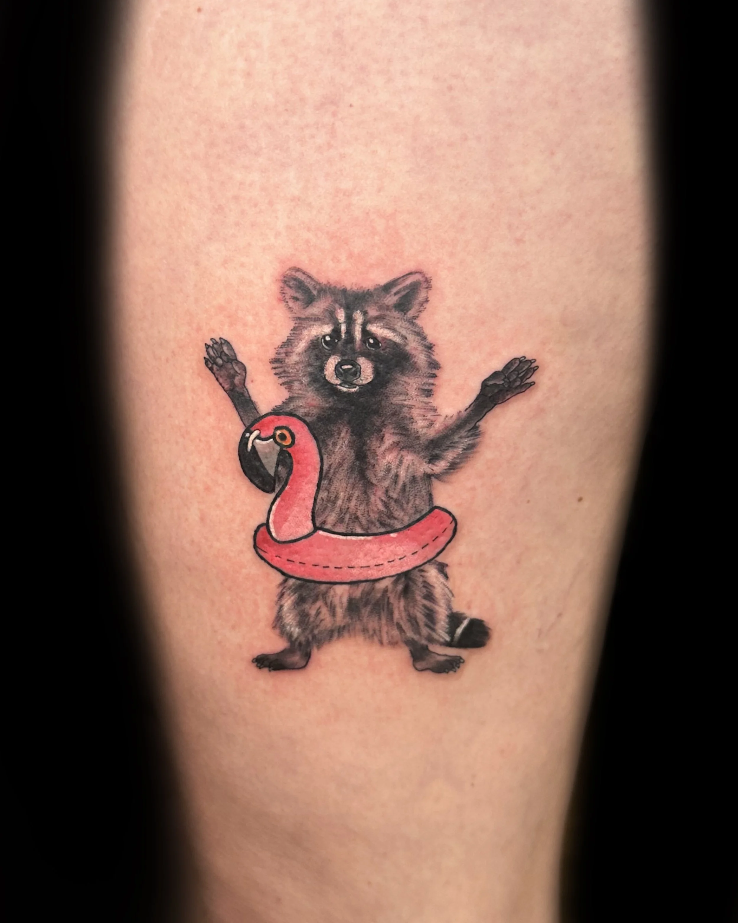 Tattoo of a raccoon with its arms raised, wearing a pink flamingo pool float around its waist.