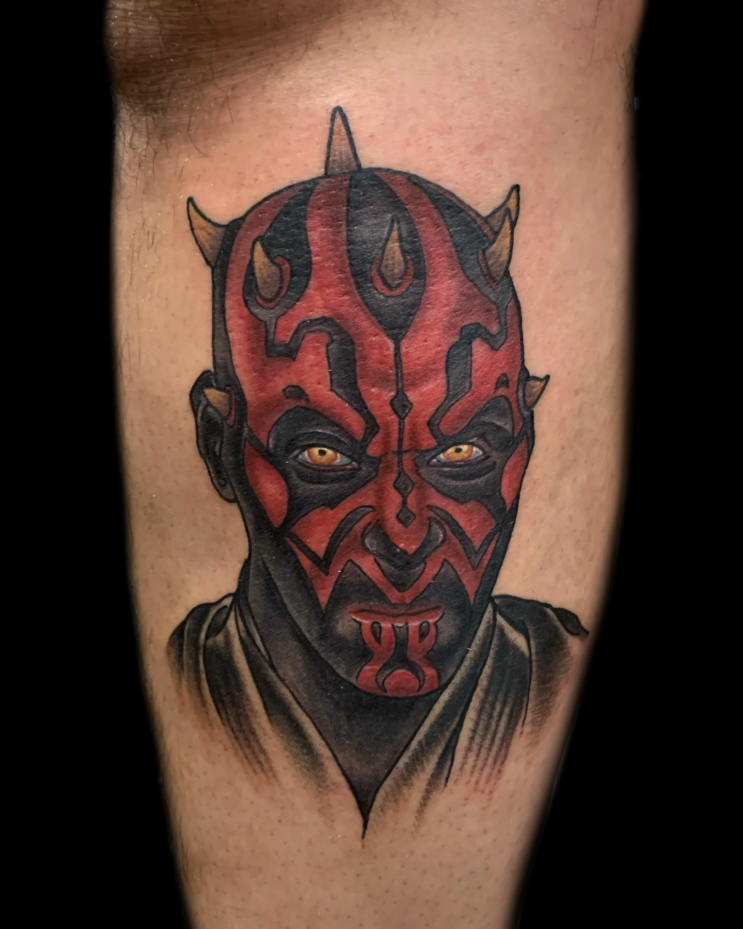 Tattoo of a person with red and black face paint resembling a demon, with yellow eyes and small horns, wearing a black cloak.