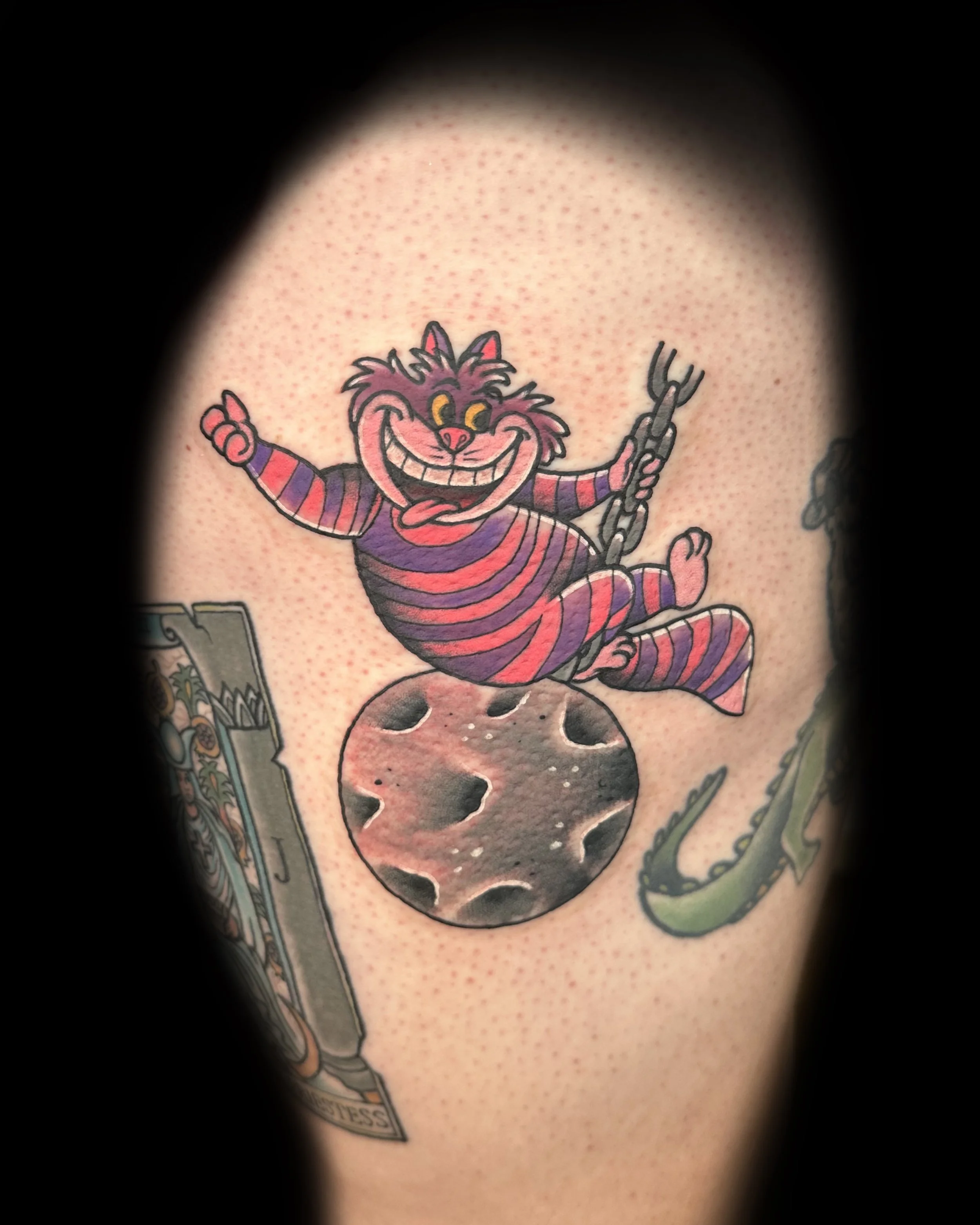 Colorful tattoo of a cartoon grinning Cheshire Cat from Alice in Wonderland, sitting on the moon with a swing hanging from its paw, and a tape measure and a snake nearby.