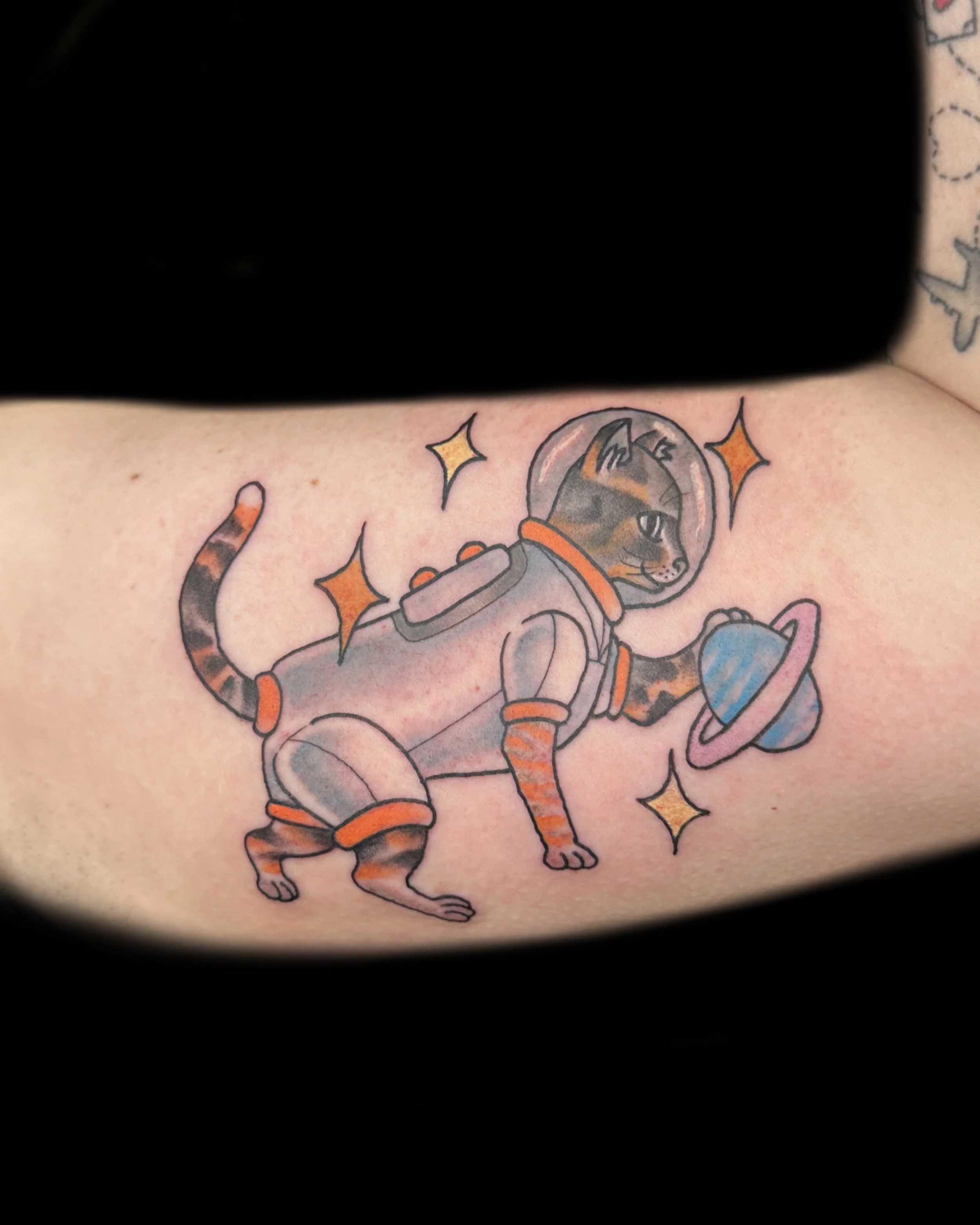 Tattoo of a cat in an astronaut suit holding a planet with a ring, surrounded by stars.