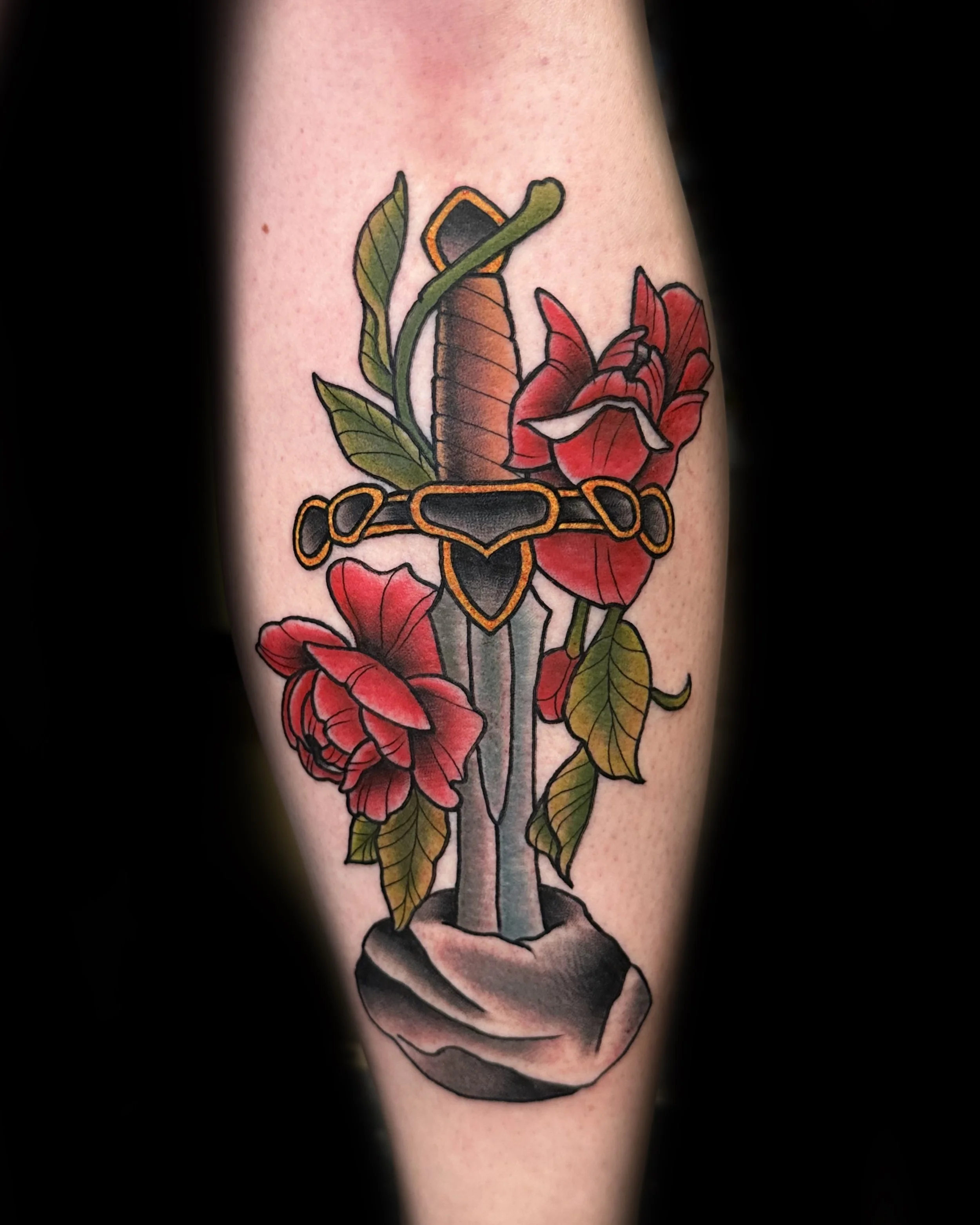 Tattoo of a sword with roses and leaves, and a black and orange dagger, on a person's arm.