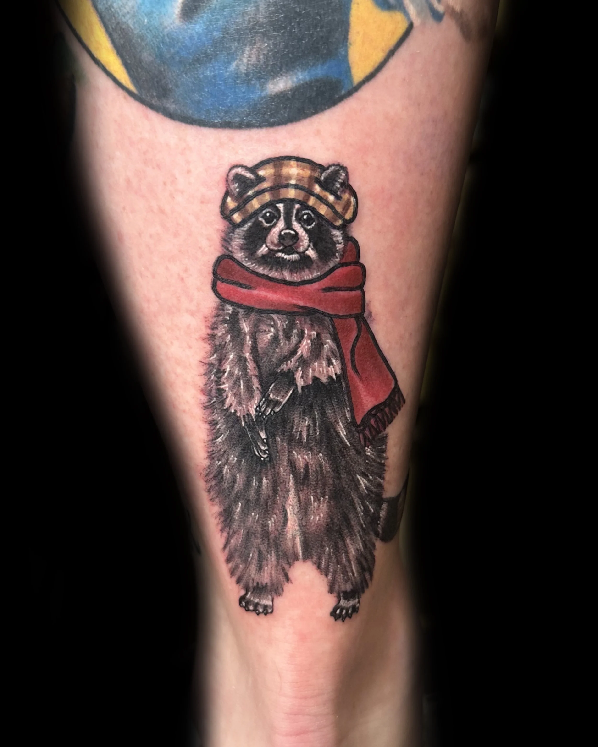 Tattoo of a raccoon wearing a cap and red scarf.