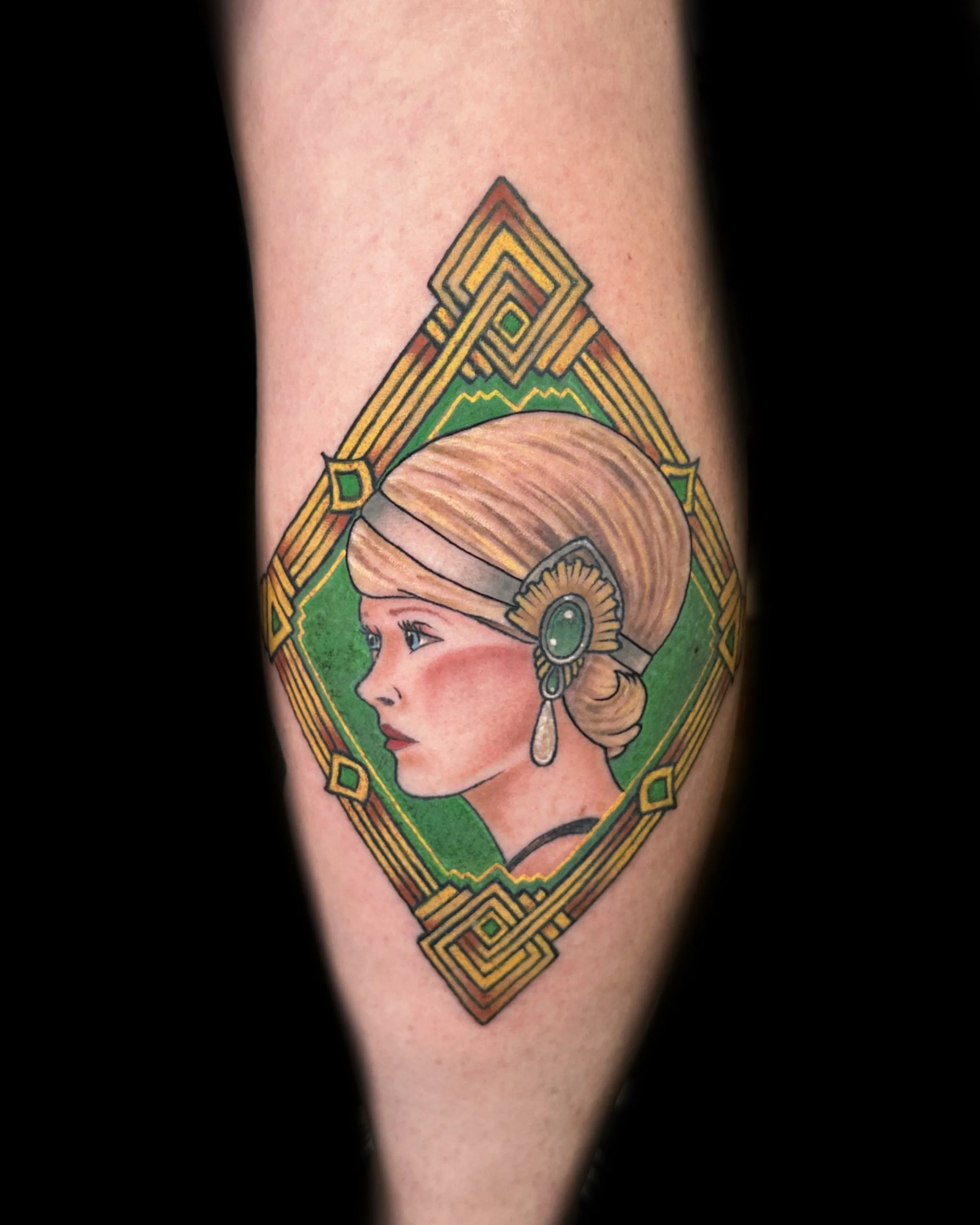Colorful tattoo of a woman in profile with vintage hairstyle, ornate hairpiece, teardrop earring, and black choker, framed by an intricate geometric border on the arm.