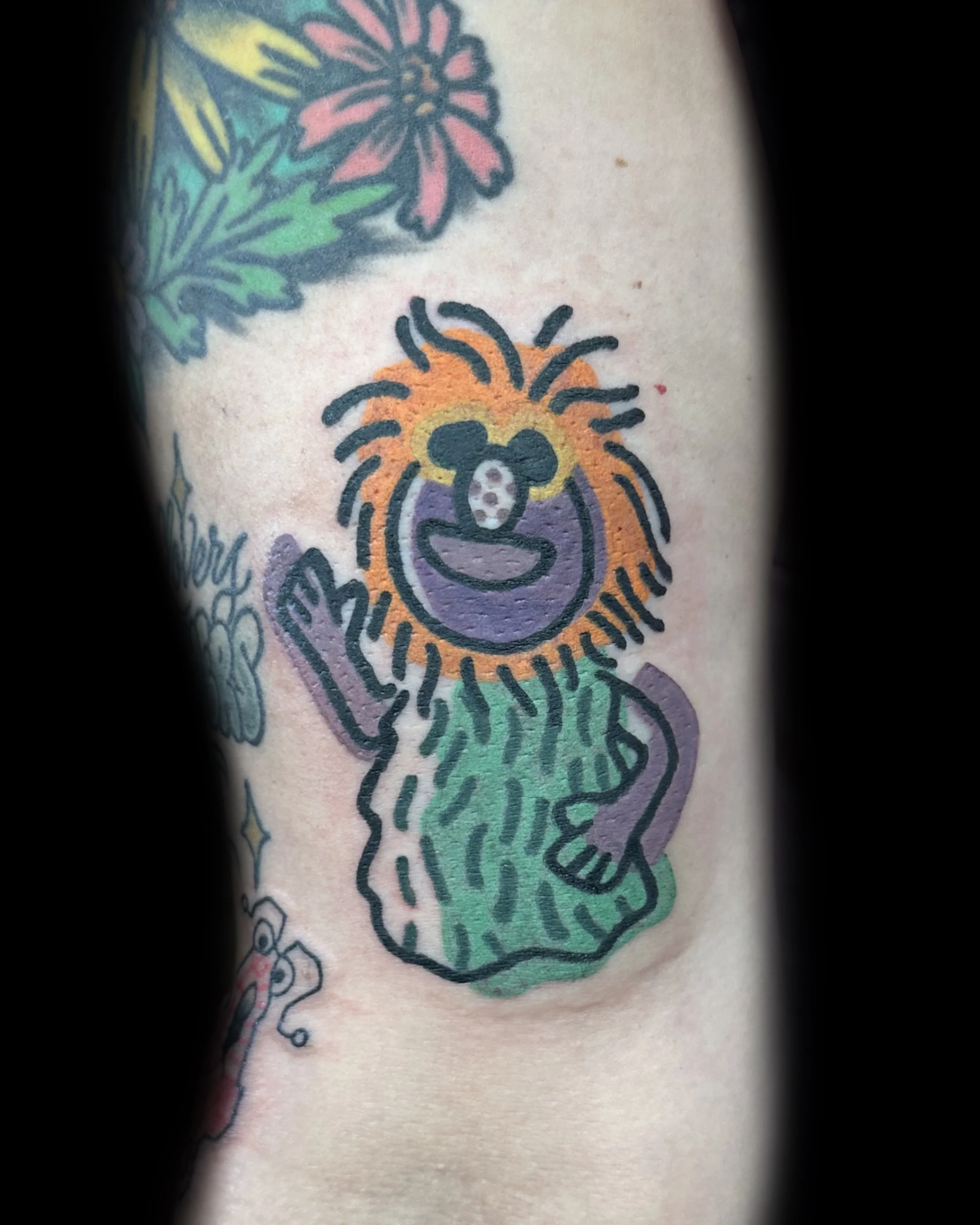 Colorful tattoo of a smiling, humanoid character with a lion's mane, waving.