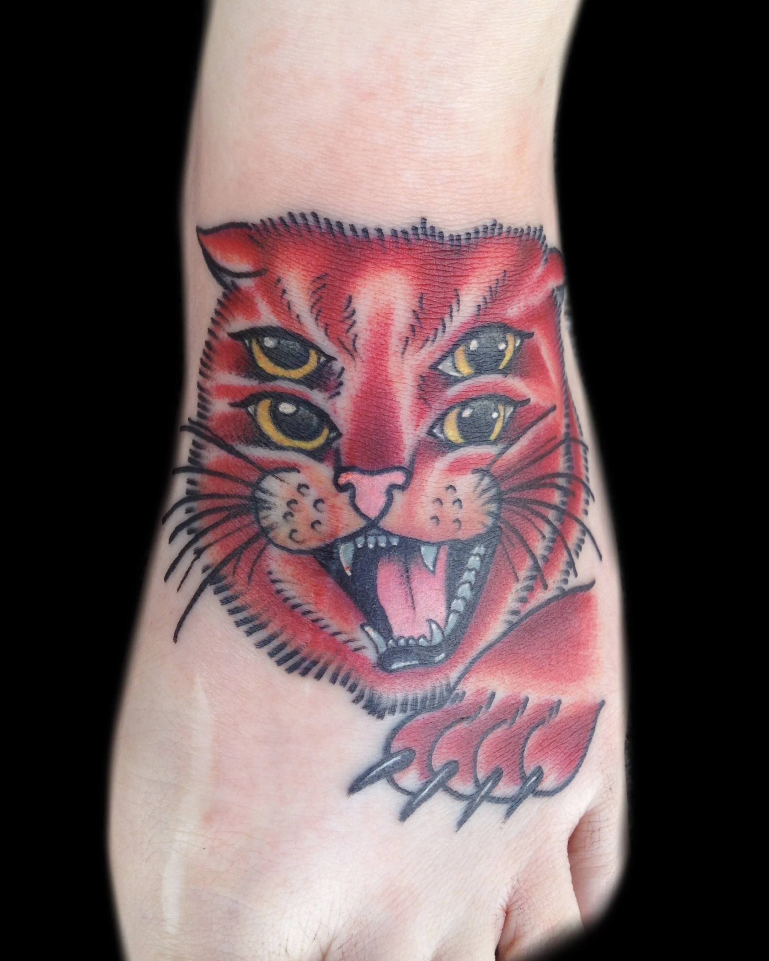 Tattoo of a red tiger face with four yellow eyes and an open mouth showing sharp teeth on a person's arm.