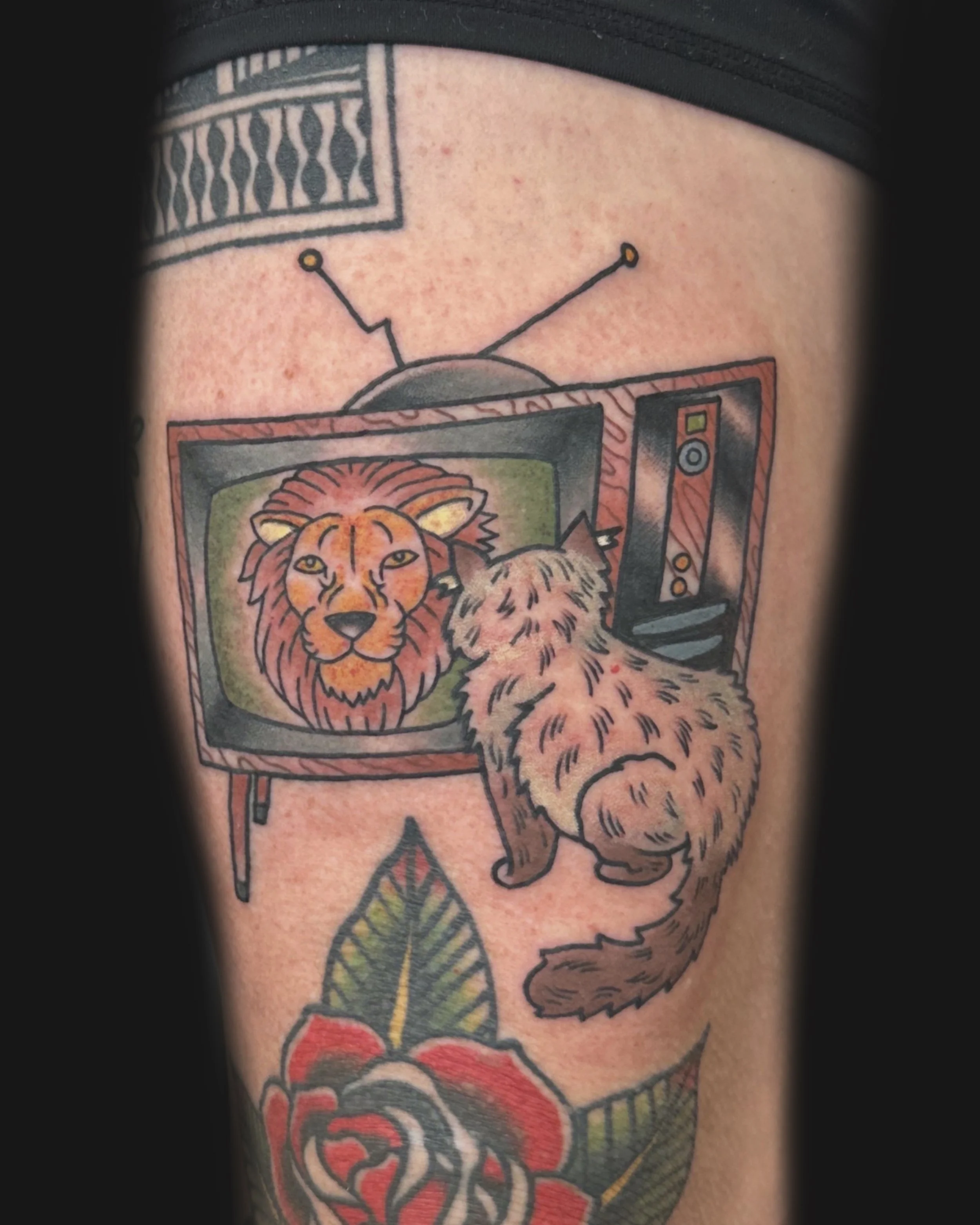 A tattoo of a cat watching a vintage television with a lion's face on the screen, and a flower below.