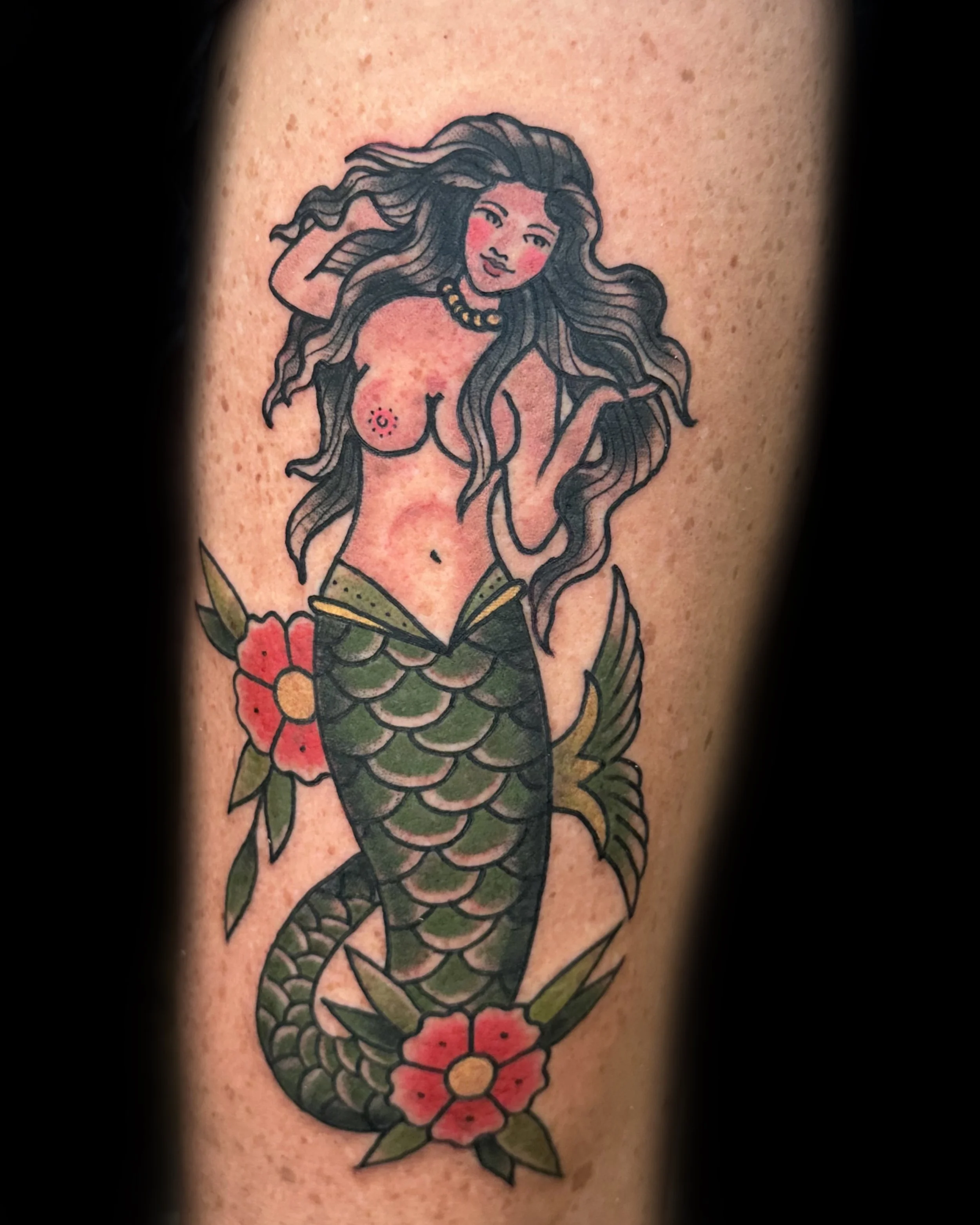 Tattoo of a mermaid with long wavy black hair, pink cheeks, and a jewelry necklace. The mermaid's upper body is topless, with visible nipples, and a belly button. She has a green-scale tail with flowers around it, and there's a small tattoo on her ri