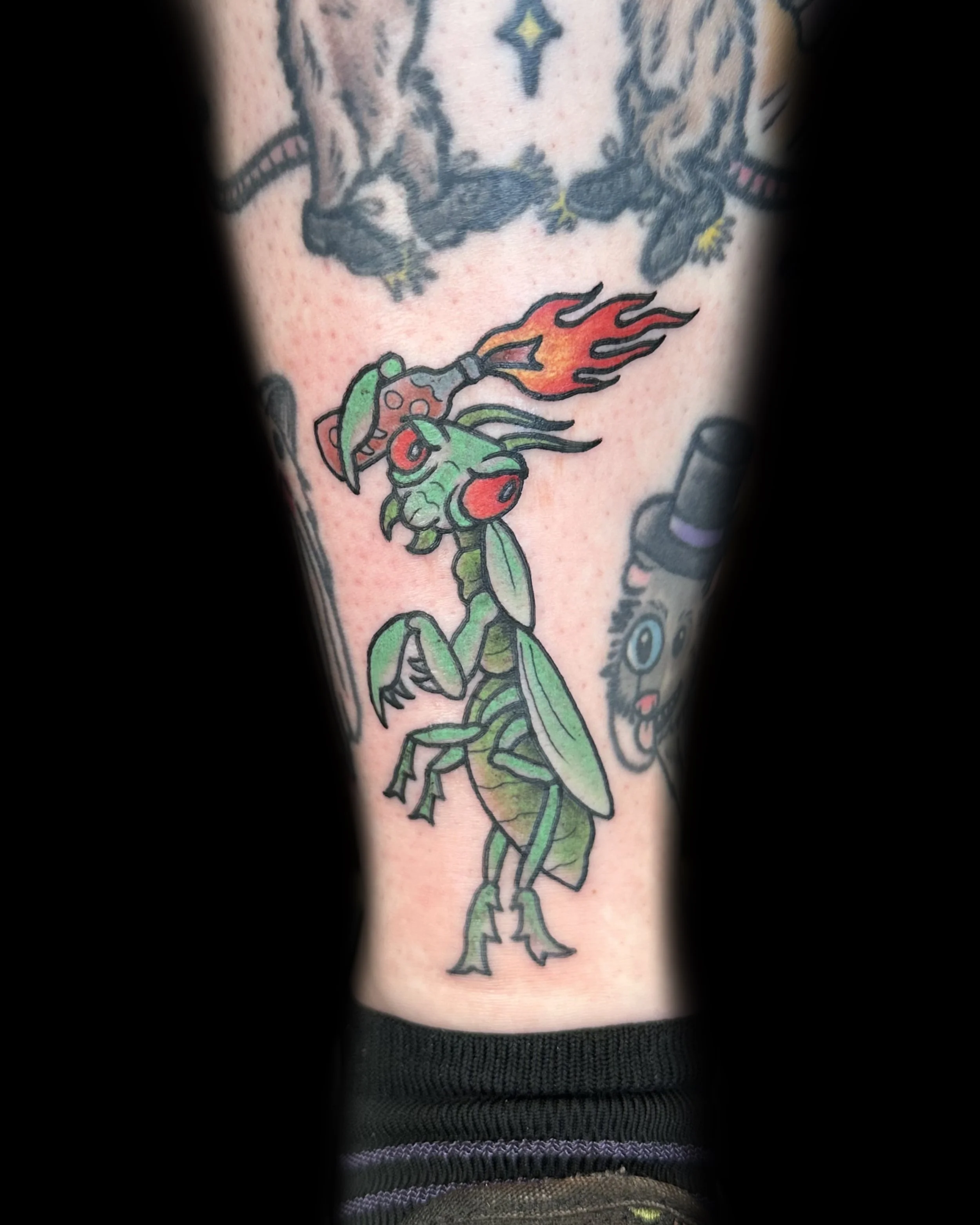 Colorful tattoo of a cartoonish praying mantis holding a flaming sword, with other tattoos partially visible around it.