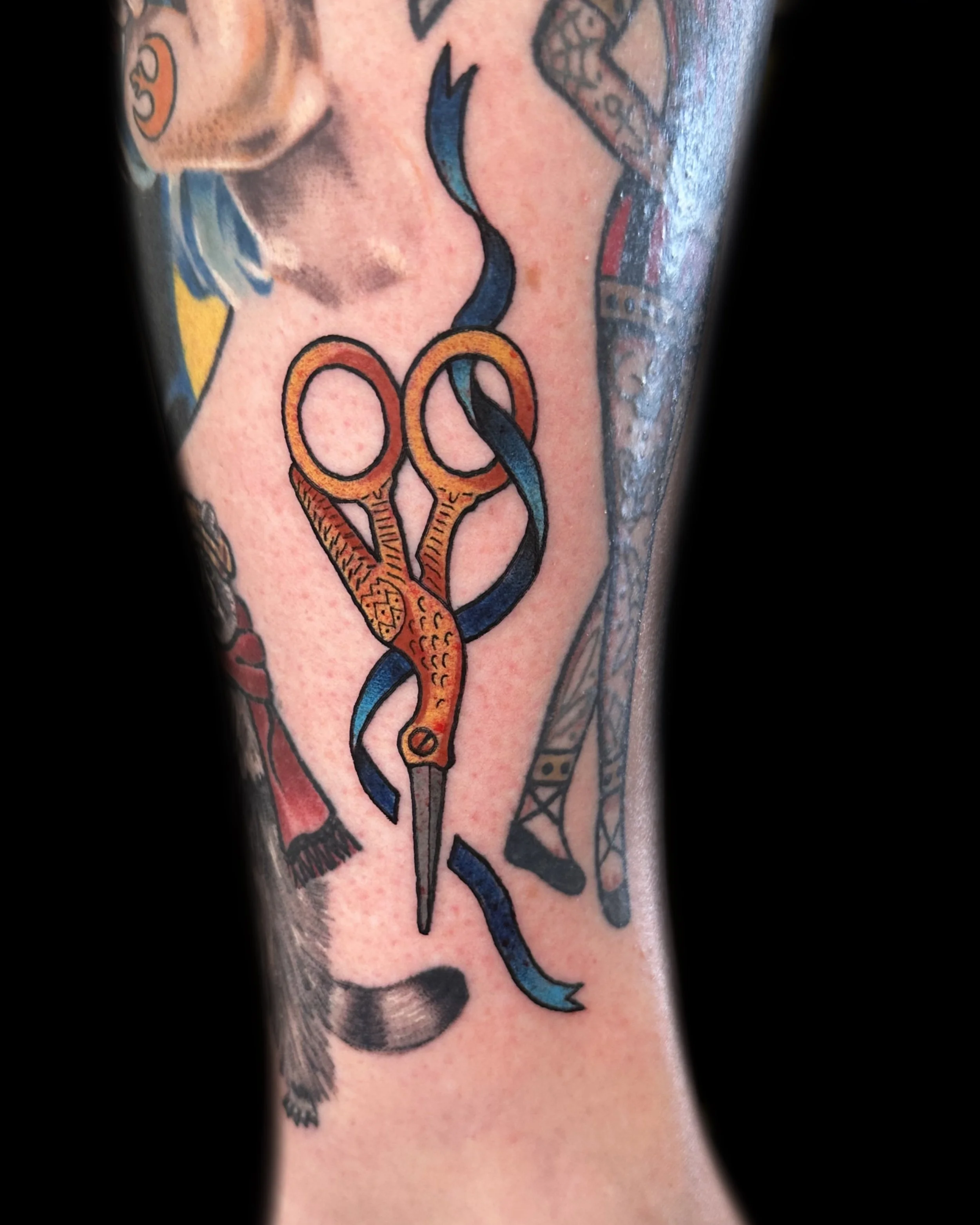 Tattoo of orange scissors with a blue ribbon intertwined, on a person's arm.