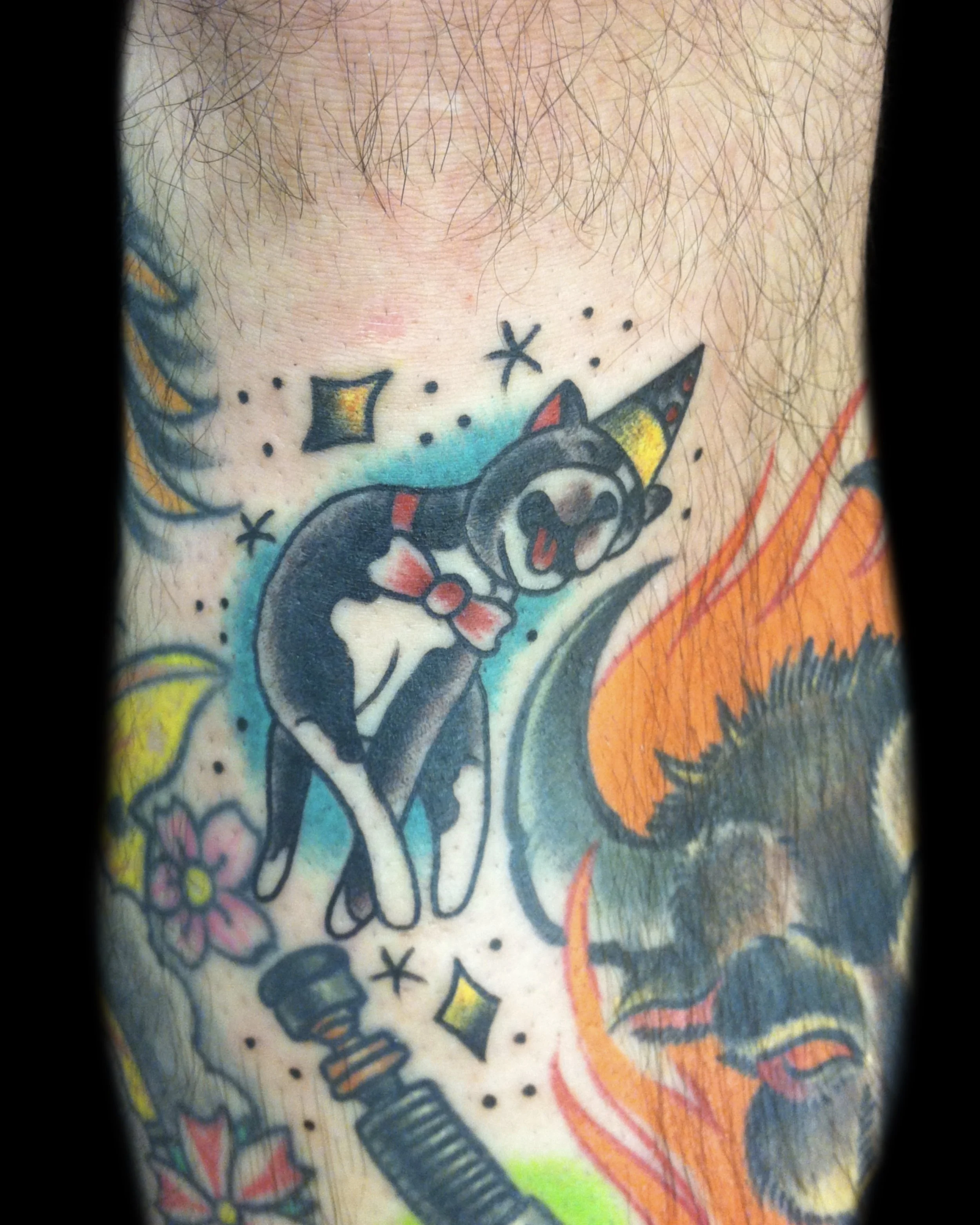 Colorful tattoo of a black and white dog with a red bow tie, sticking out its tongue, surrounded by stars, diamonds, and colorful elements on skin with body hair visible.