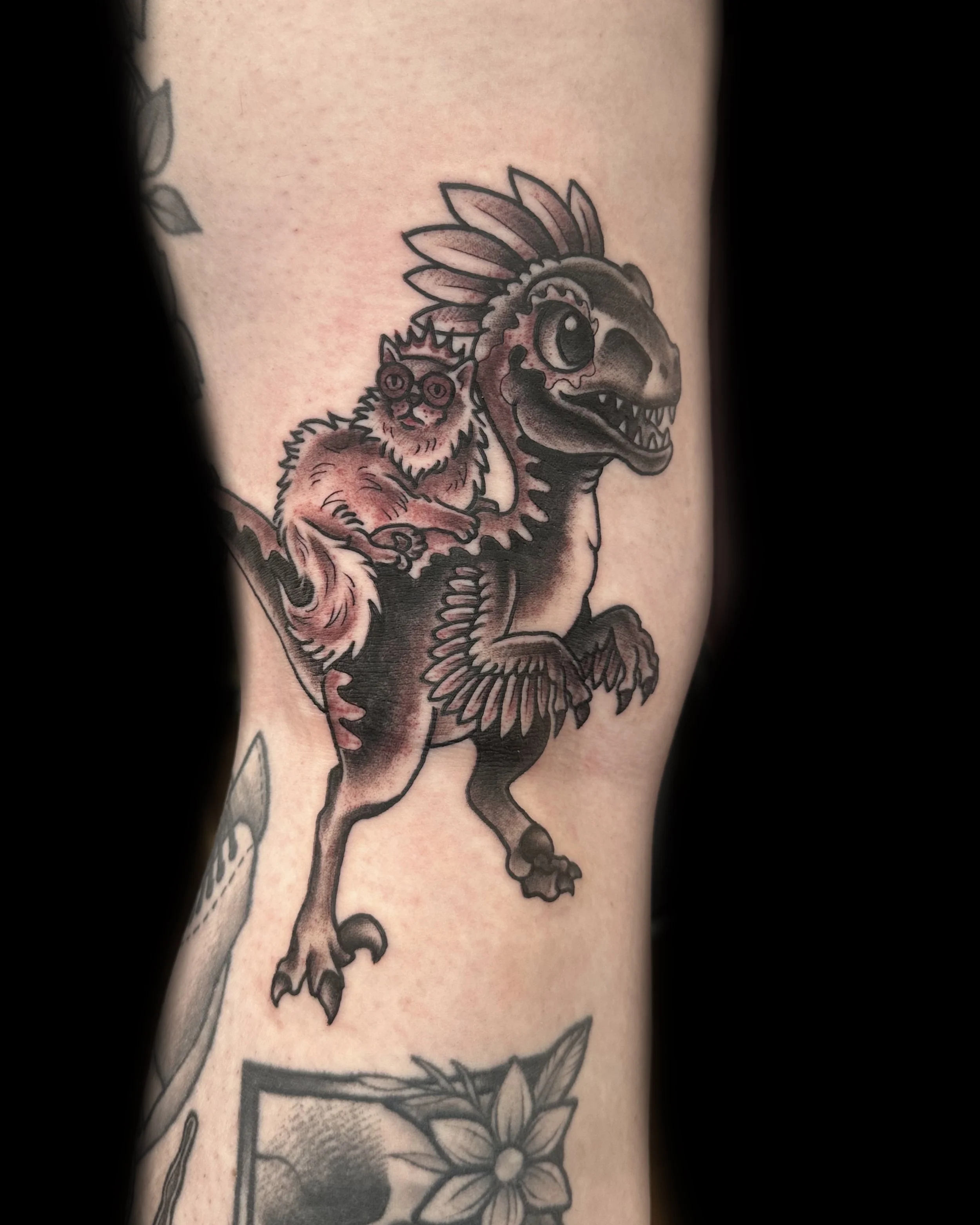 Tattoo of a cartoon dinosaur with a feathered crest on its head, carrying a small cat wearing glasses and a crown.