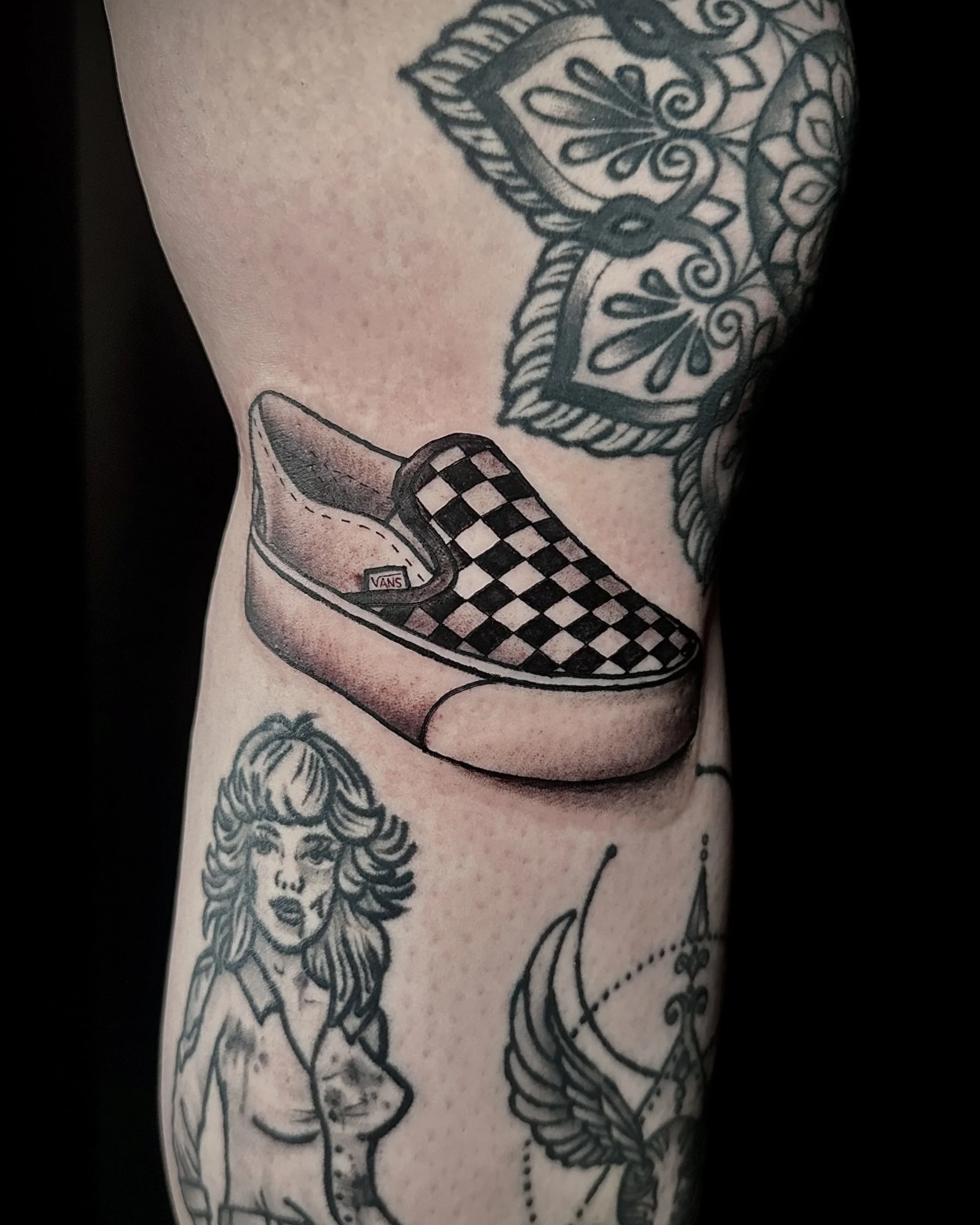 Tattoo of a Vans checkered slip-on shoe on a person's arm, along with partial inked images of a woman with curly hair and a tattooed figure with a winged design.