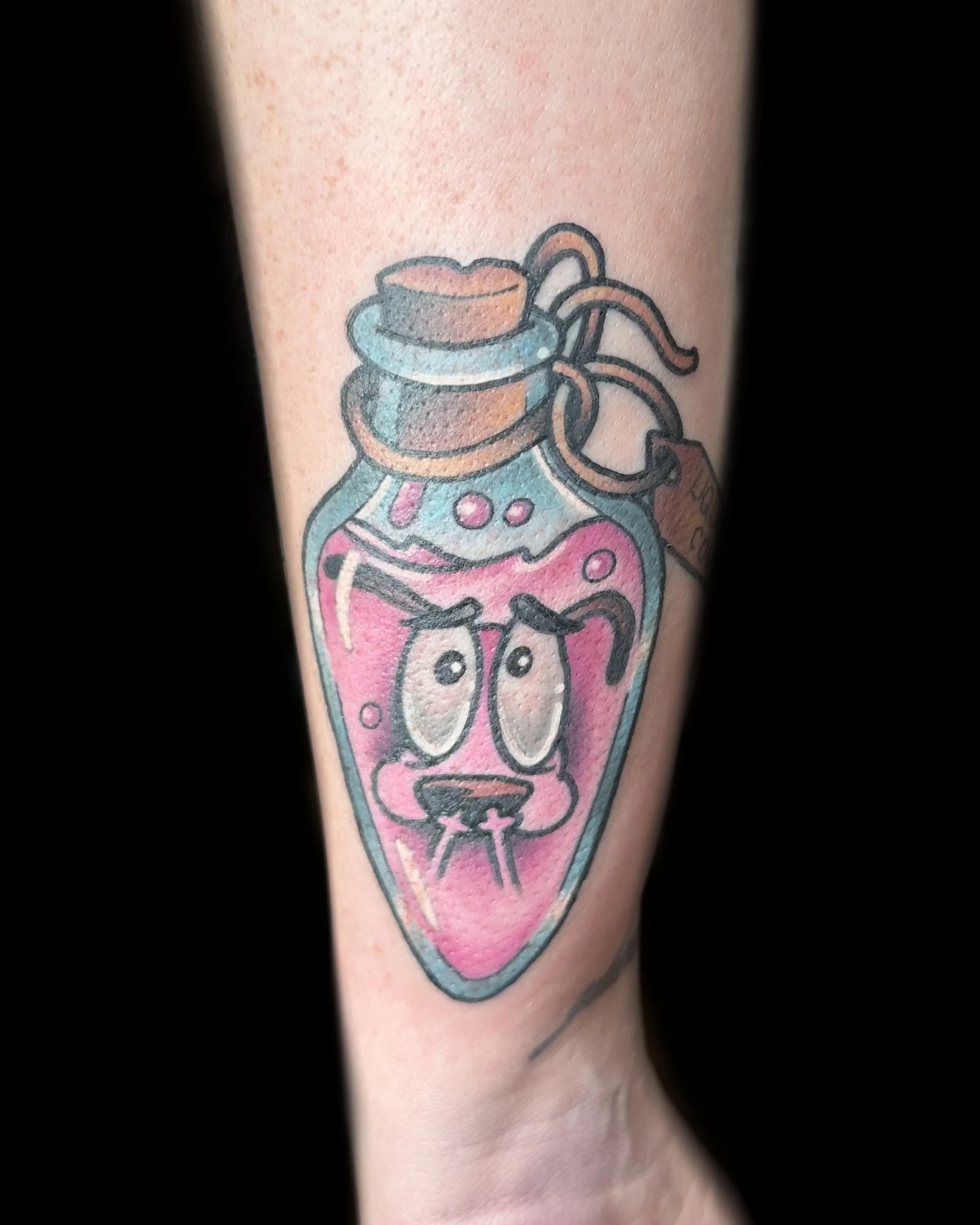 Colorful tattoo of a cartoon heart with human-like eyes and an expressive face, in a pink and teal color scheme, with a small tag attached.