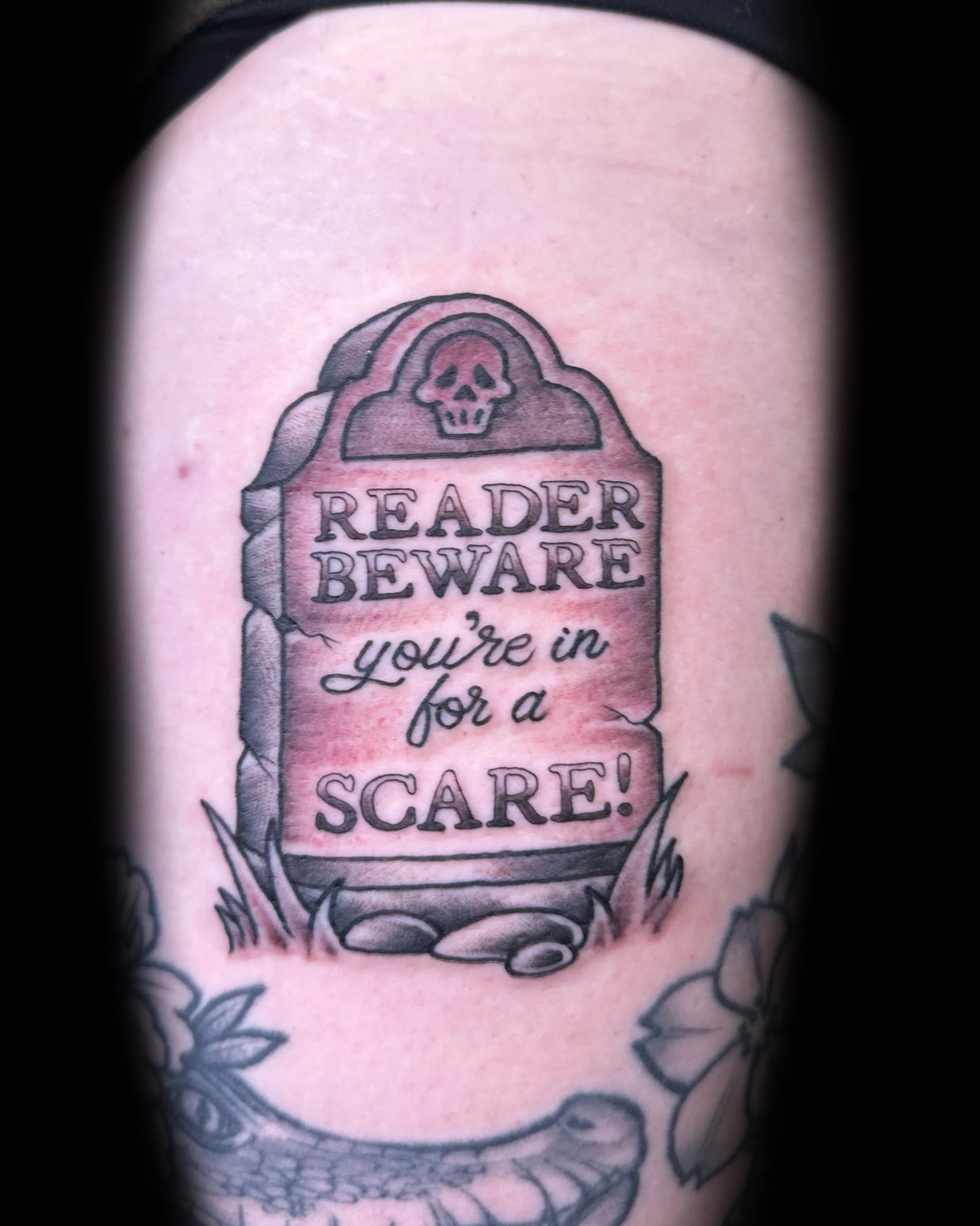 Tattoo of a tombstone with a skull at the top, the words "READER BEWARE" and "you're in for a SCARE!" written on it, surrounded by flowers and rocks.