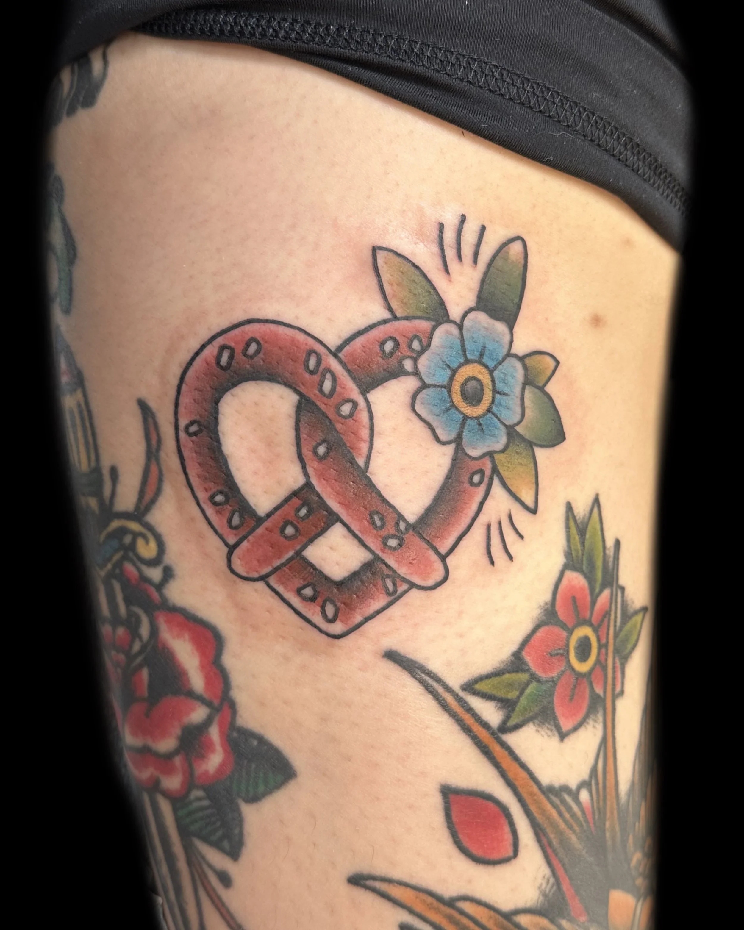 Tattoo of a heart-shaped pretzel with a blue flower and green leaves behind it.