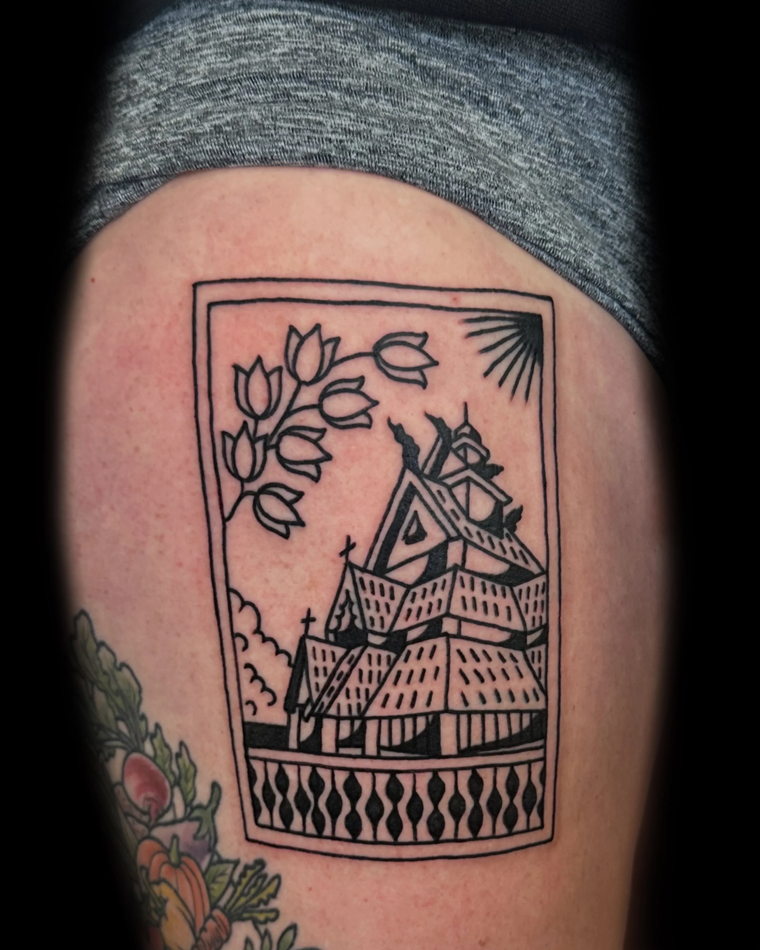 Black outline tattoo of a traditional-style house with a steep roof, steeples, and a railing, surrounded by a rectangular border. A branch with flowers and leaves extends from the top left corner, and a sunburst is in the top right corner.