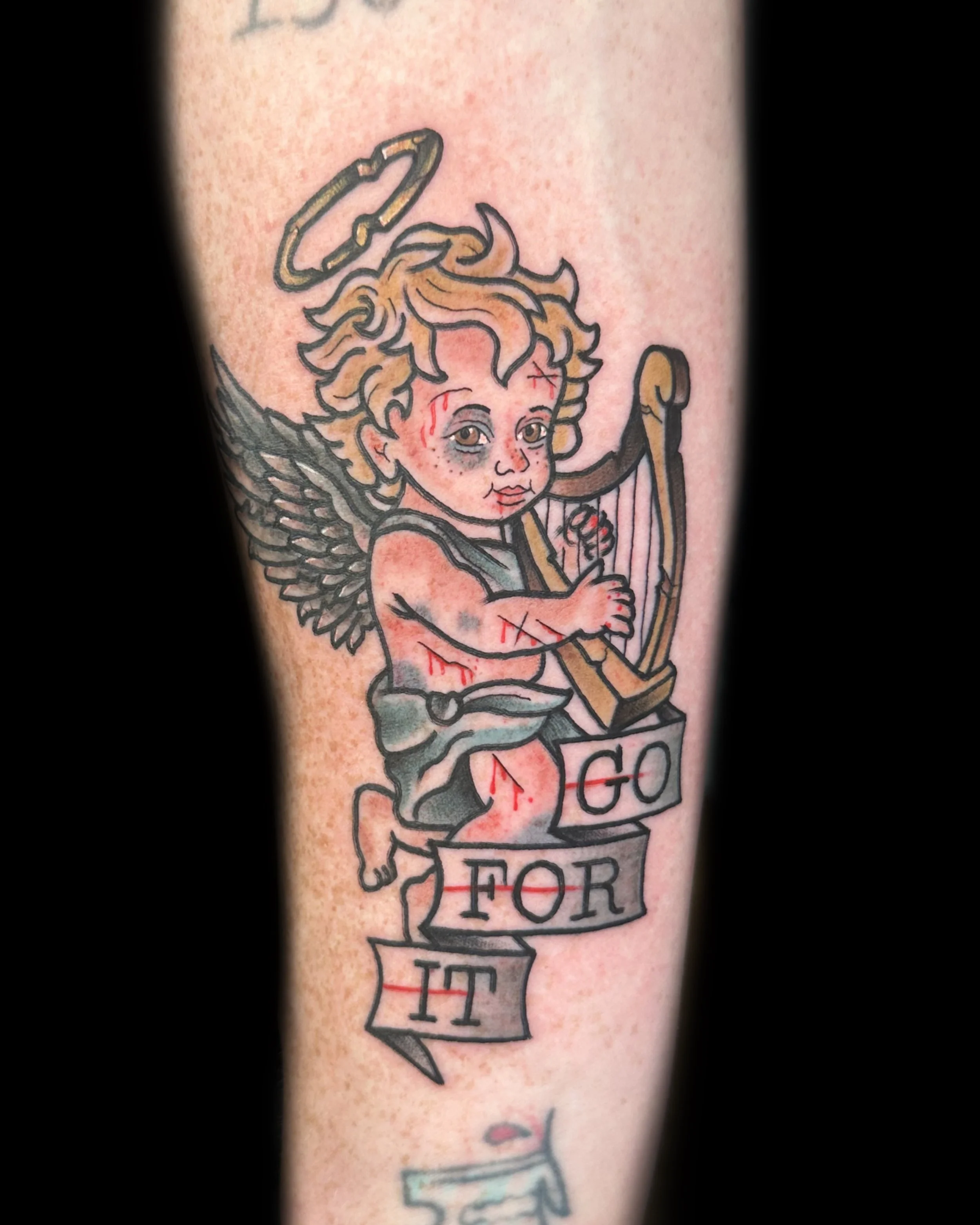 Colorful tattoo of a cherub with wings, a halo, and a bow, holding an arrow, with a banner that reads 'GO FOR IT'.