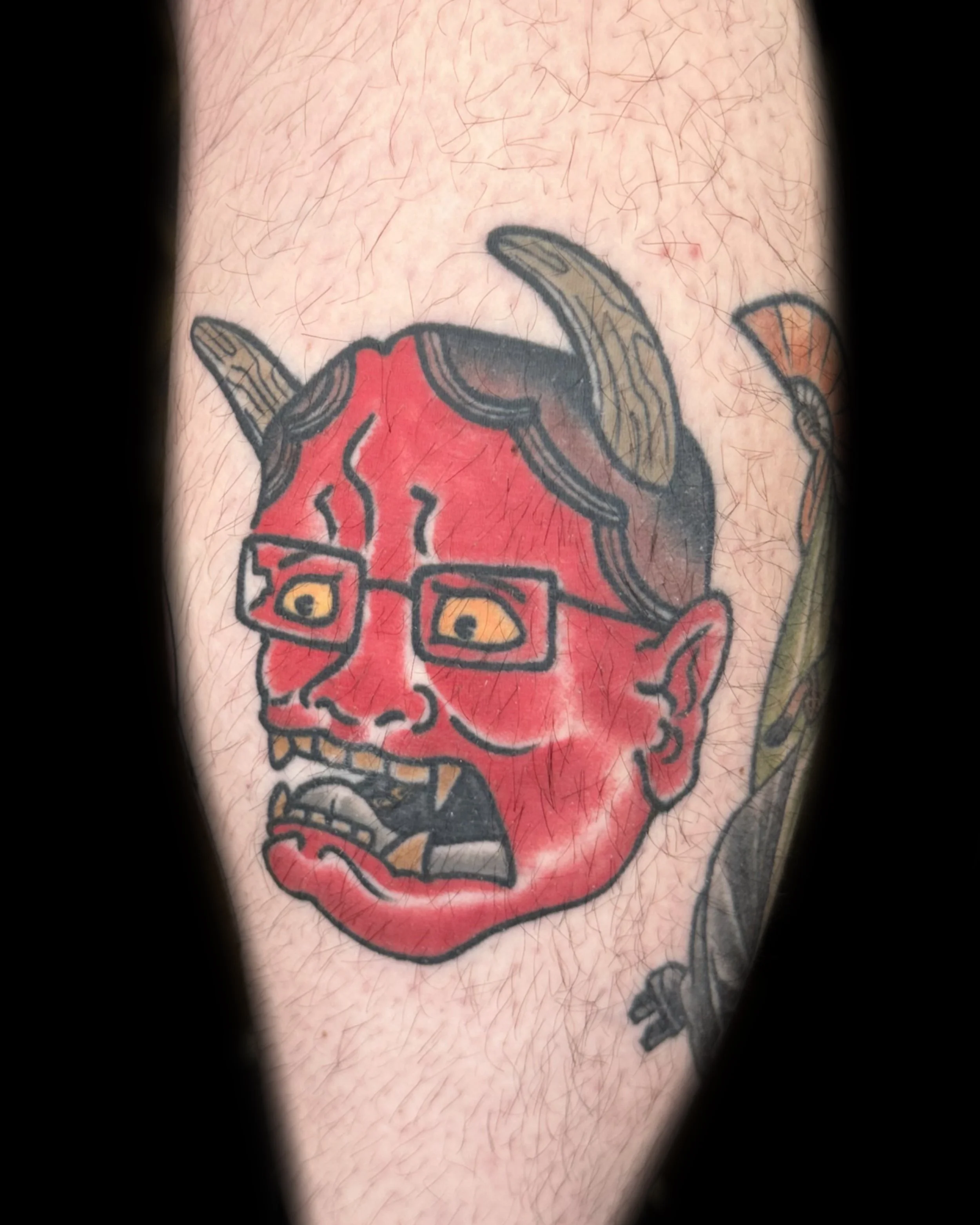 Tattoo of a cartoonish red demon face with glasses, sharp teeth, and a mischievous expression.