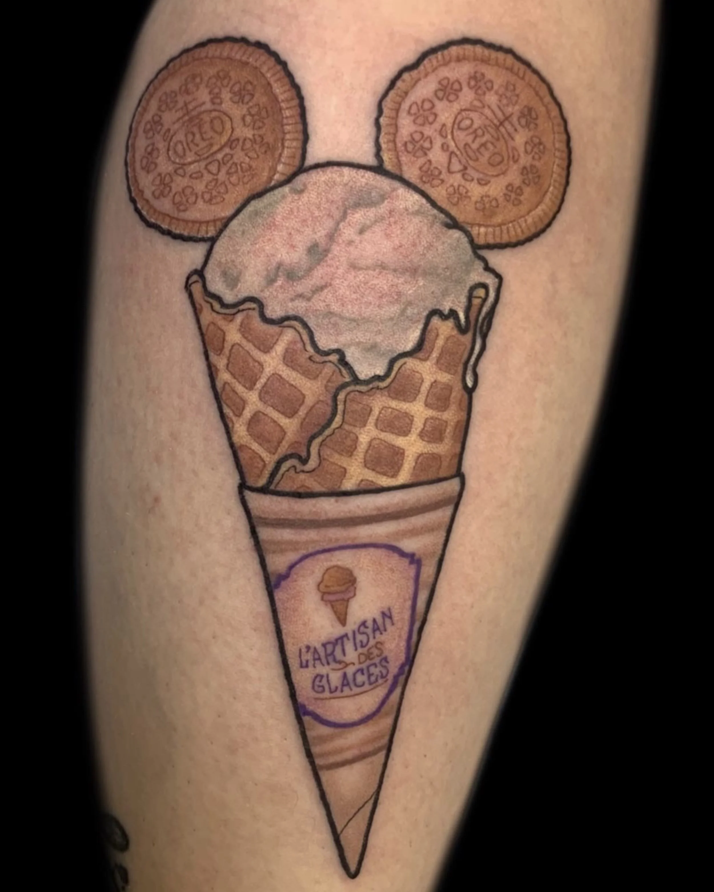 Tattoo of an ice cream cone with two cookies resembling Oreo cookies and the text 'L'ARTISAN DES GLACES' on the cone, with a logo of a small ice cream cone above the text.