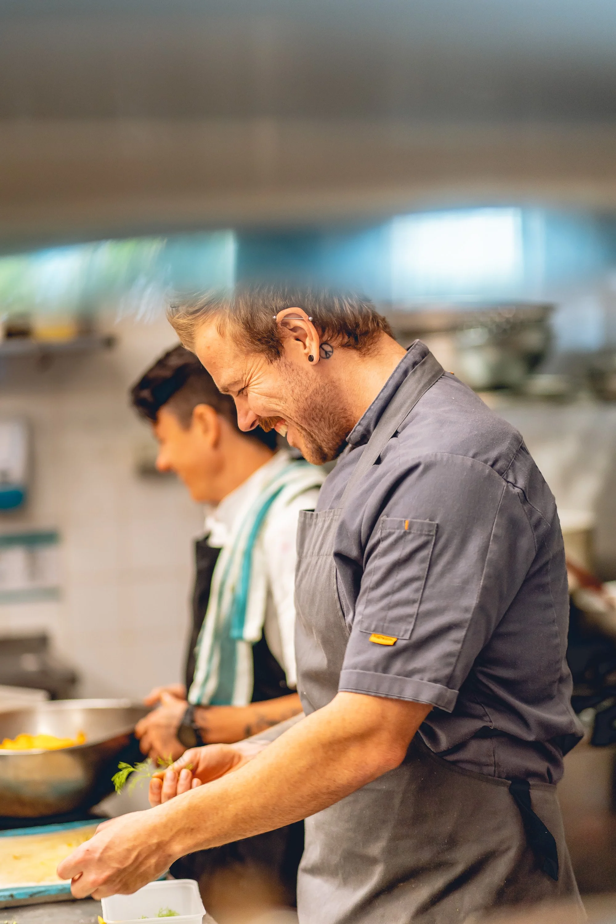 Two chefs in a commercial kitchen preparing food, one with long hair and the other with short dark hair, both wearing aprons.