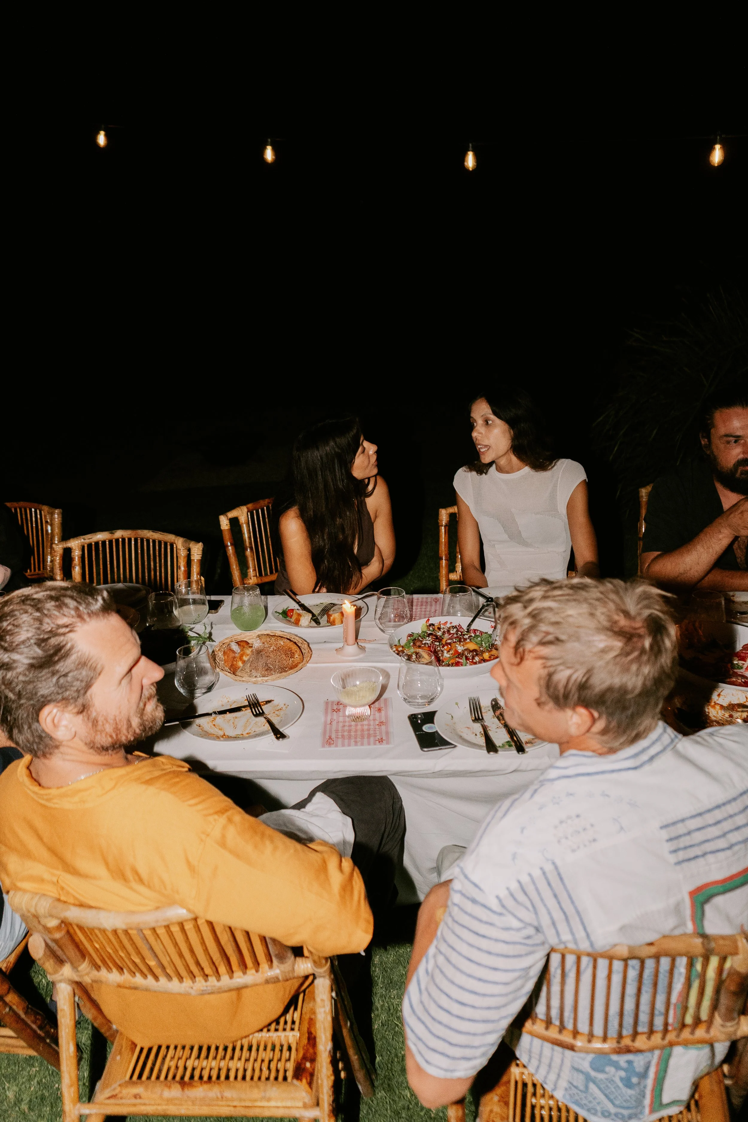 People sitting at a dinner table outdoors during the night, engaged in conversation, with food and drinks on the table and string lights hanging above.