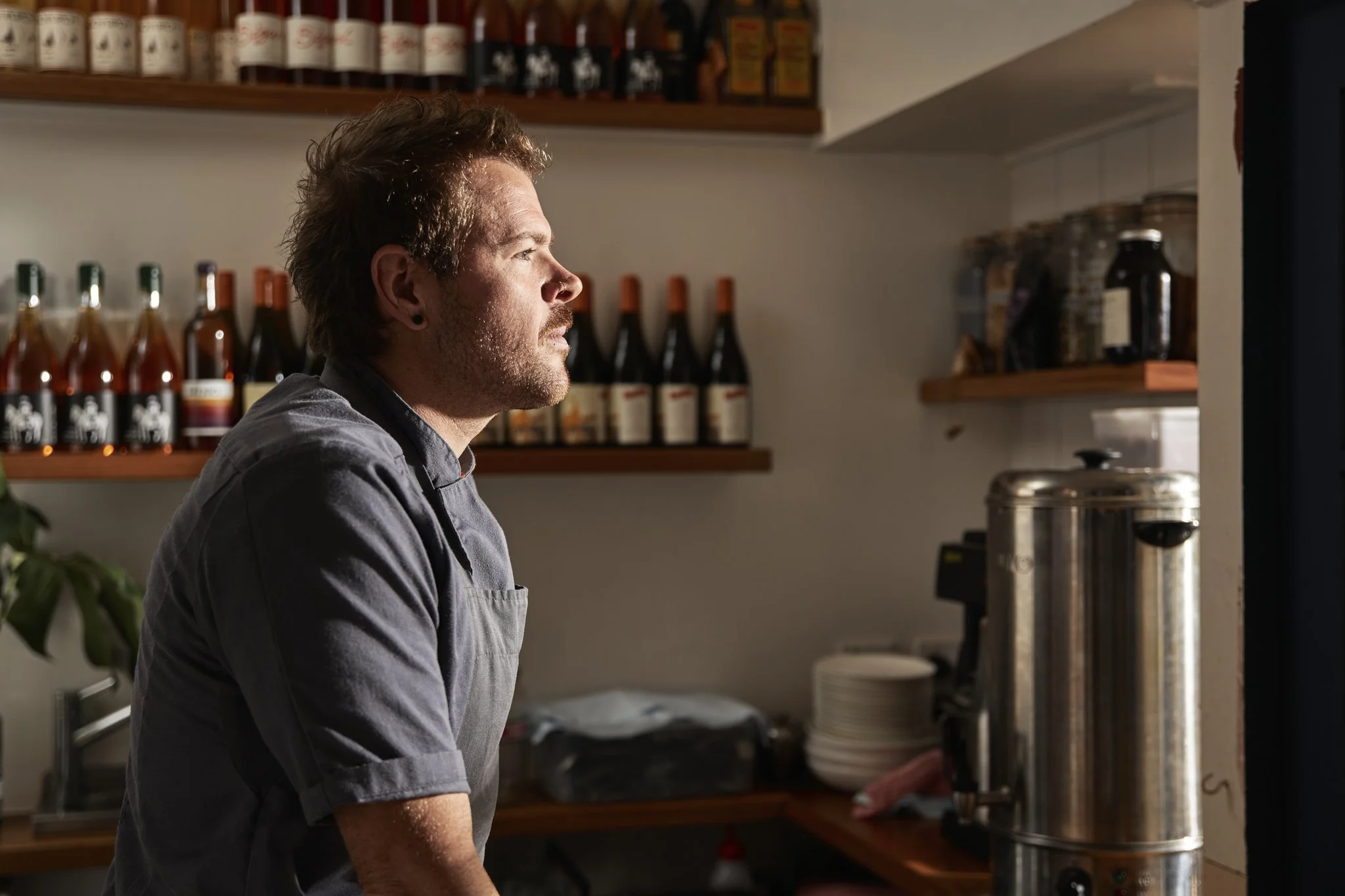 A man with short, messy hair, wearing a gray shirt and apron, standing in a kitchen. He is looking to his right, next to shelves with bottles and jars of spices, sauces, and other ingredients. There is a large stainless steel coffee urn on the counter. The scene appears to be in a cozy restaurant or cafe kitchen.