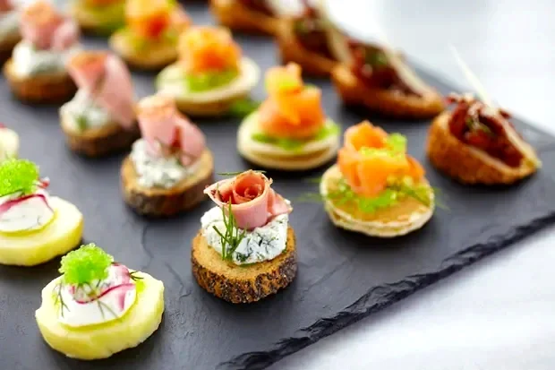 Assorted canapés on a slate serving board, topped with various meats, cheeses, and vegetables.