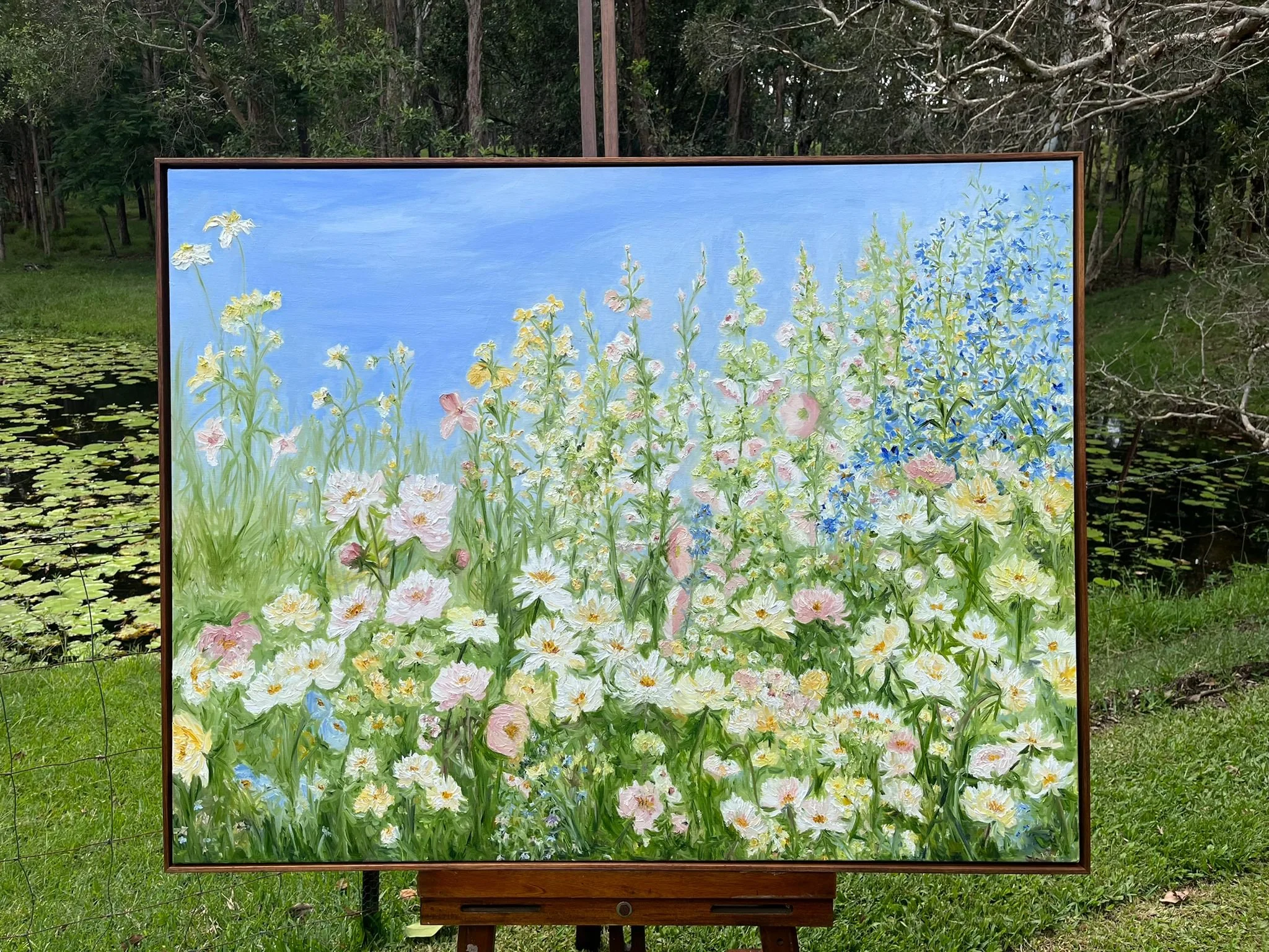 A painting of a colorful floral garden with various flowers and a blue sky in the background, displayed outdoors near a pond with lily pads.