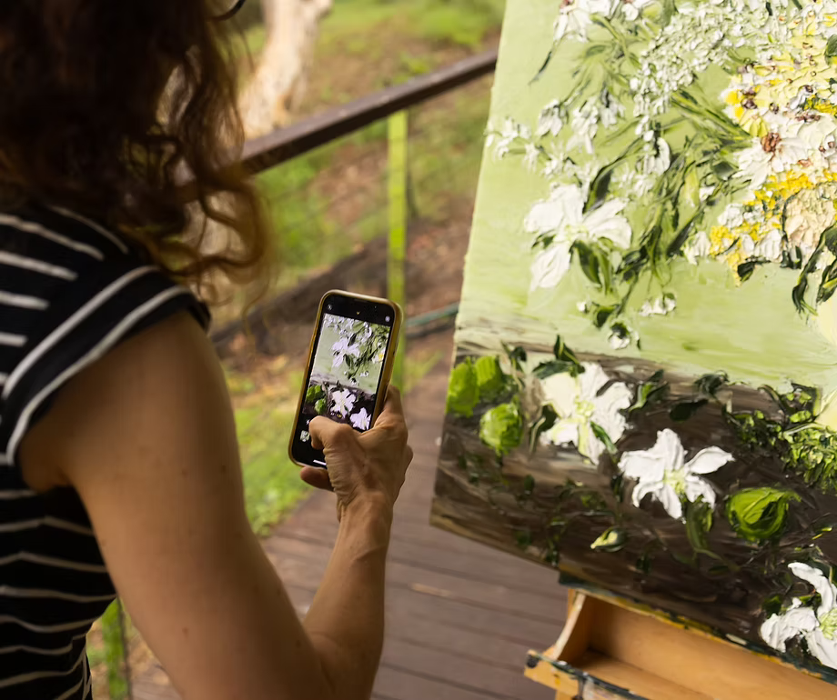 A woman taking a photo of a floral painting on an easel with her phone in an outdoor setting.
