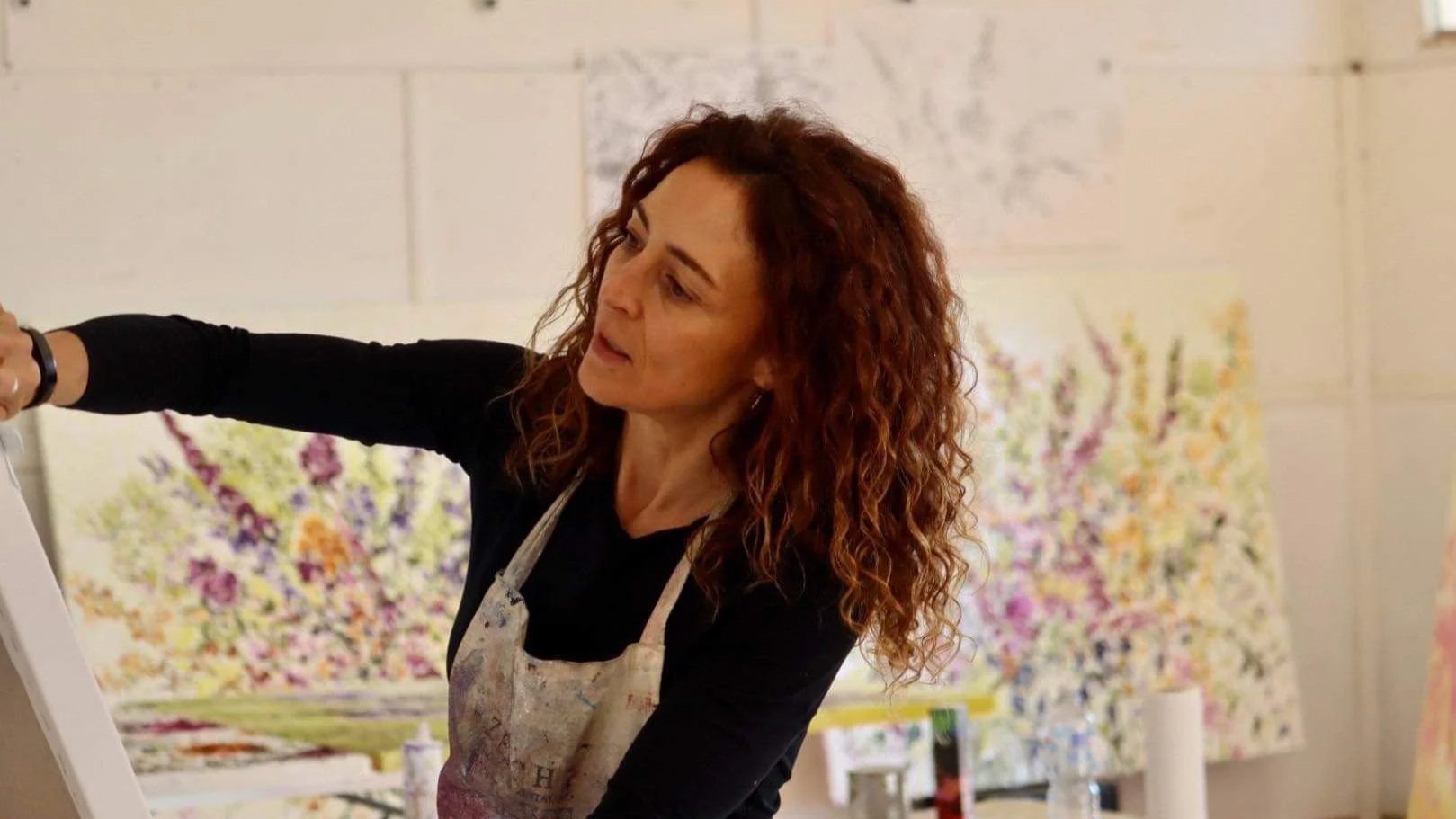 A woman with curly red hair wearing a black shirt and apron, painting on a canvas in an art studio with colorful floral paintings in the background.